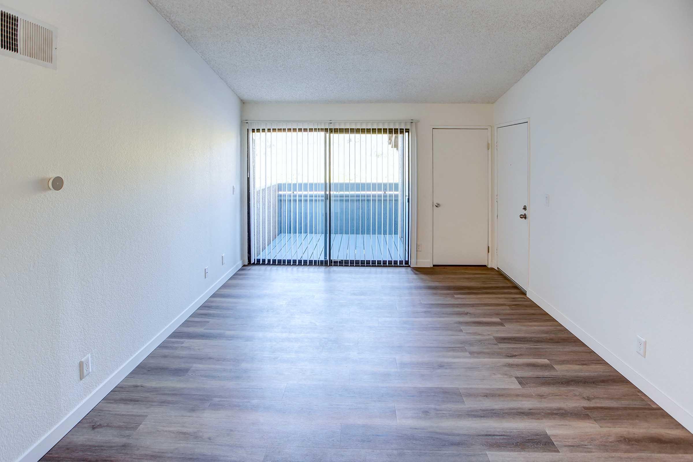 Bright and empty living space with light-colored walls, a wooden floor, and a sliding glass door featuring vertical blinds that opens to a balcony. There is a closed door on the right side, and the space appears clean and well-lit, ready for furnishing.