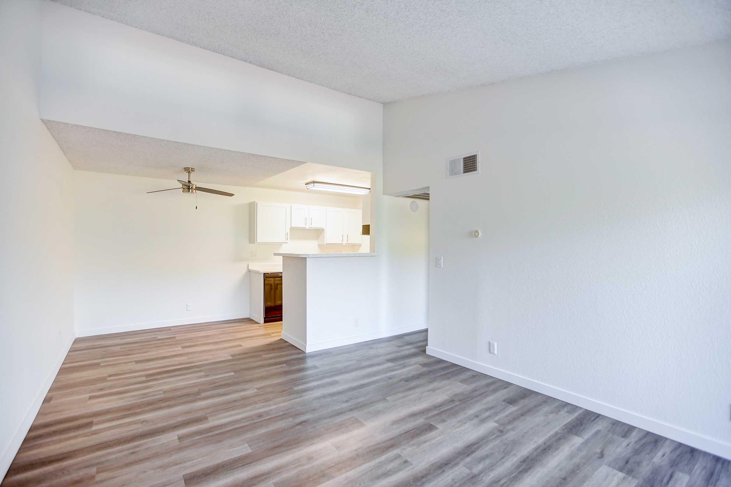 A spacious, well-lit room featuring light-colored walls and a modern, open design. The floor is covered with laminate wood, and there is a ceiling fan. An adjacent kitchen area is visible with white cabinetry.