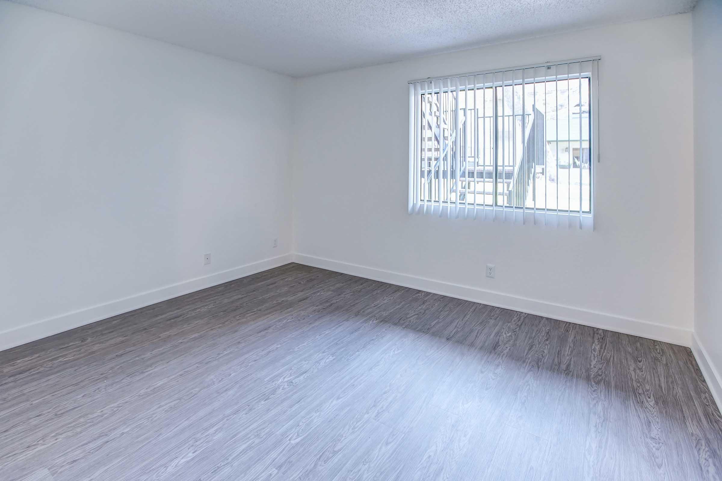 An empty room with light-colored walls and a large window featuring vertical blinds. The flooring is a light wood-like texture, and natural light is coming in through the window, creating a bright atmosphere. There are no furnishings or decorations present in the space.