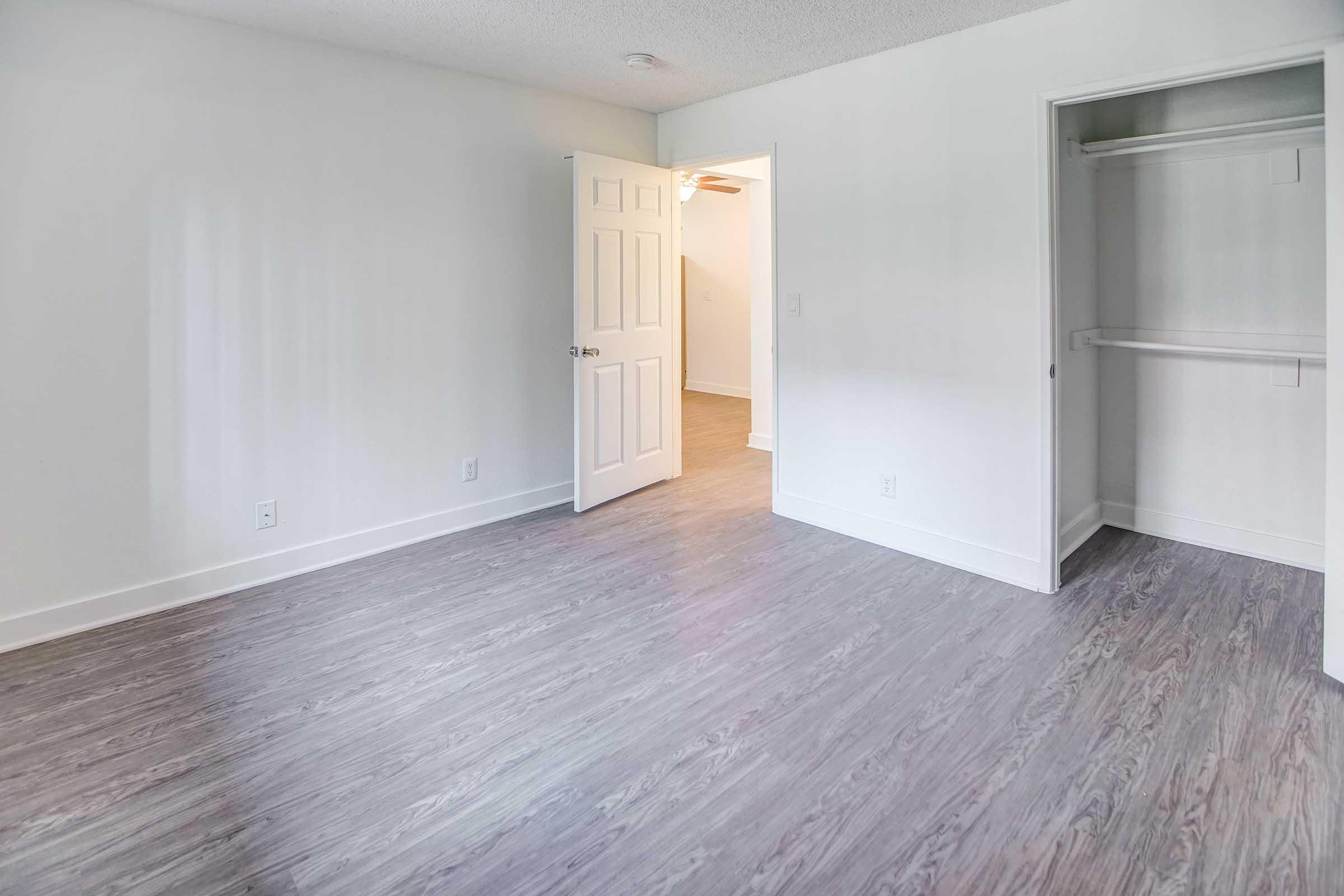 A vacant room with light gray walls and wooden flooring. There's an open door leading to another room, and a closet with sliding doors on the right. The space is well-lit, showcasing a clean and minimalist design, ideal for potential decoration or furniture arrangement.