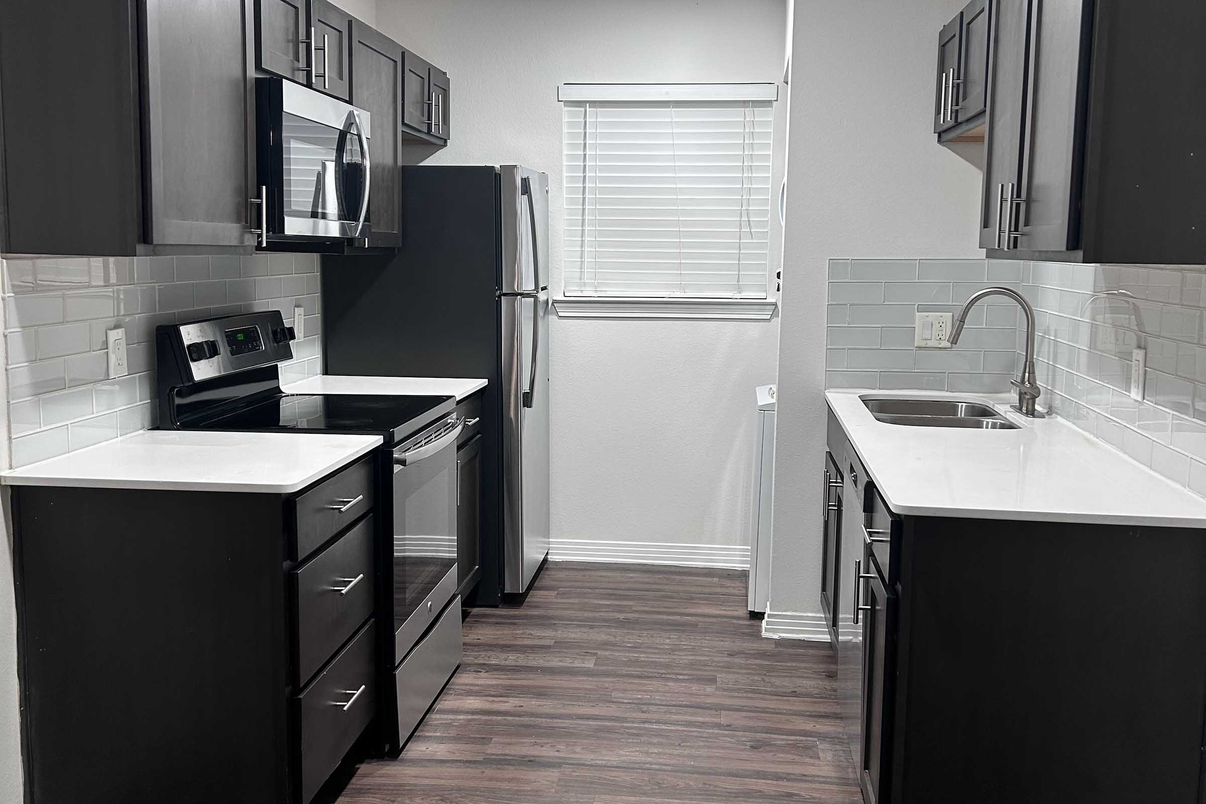 Modern kitchen featuring dark wood cabinets, stainless steel appliances including a microwave, oven, and refrigerator. The countertop is white with a matching backsplash. There is a double sink and large window with blinds, illuminating the space. The flooring is dark wood laminate.