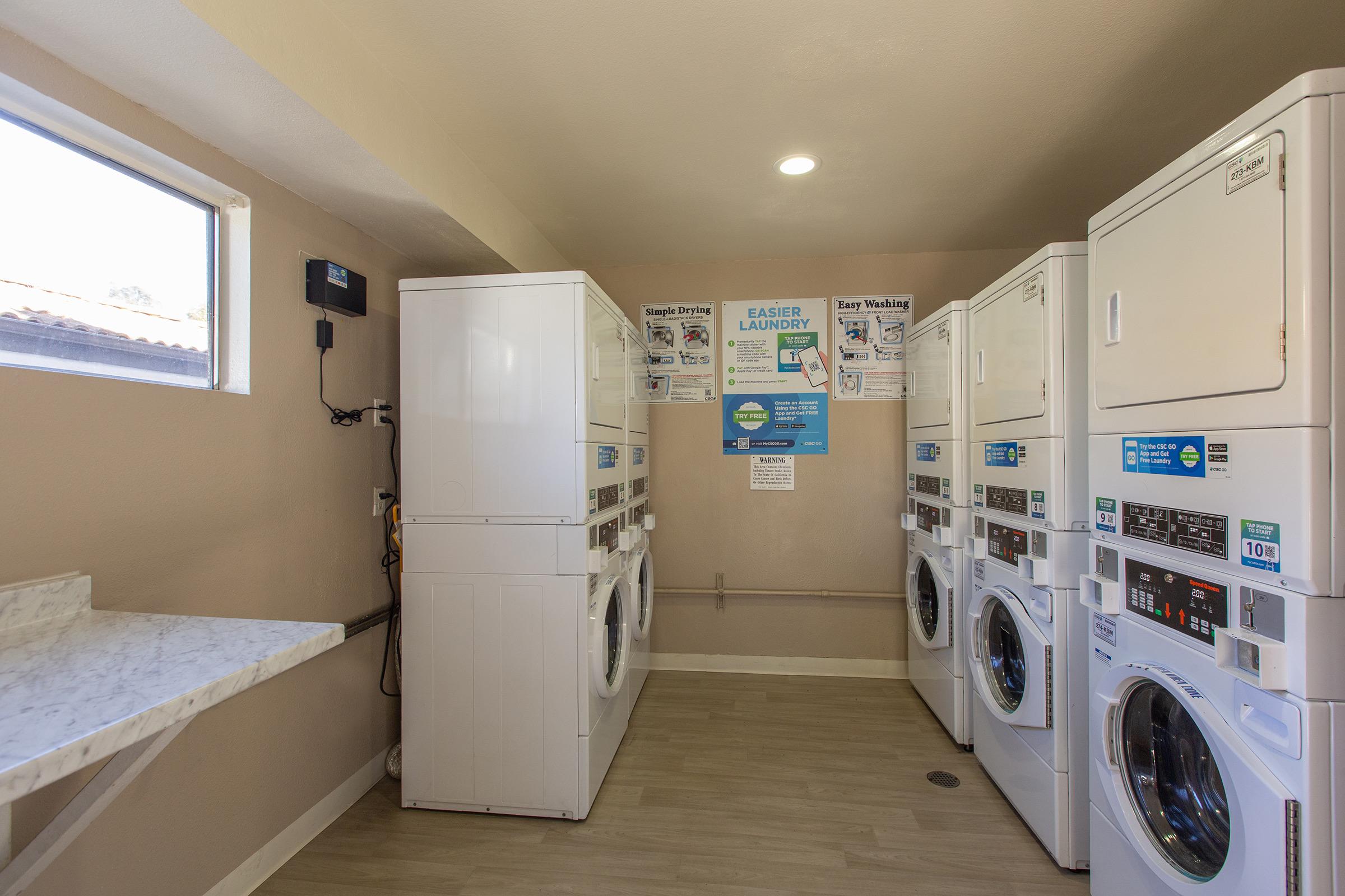 A clean laundry room featuring several white washers and dryers, with a countertop and a window for natural light. Informational posters about laundry services are displayed on the walls. The floor is light-colored, creating a bright and welcoming atmosphere.
