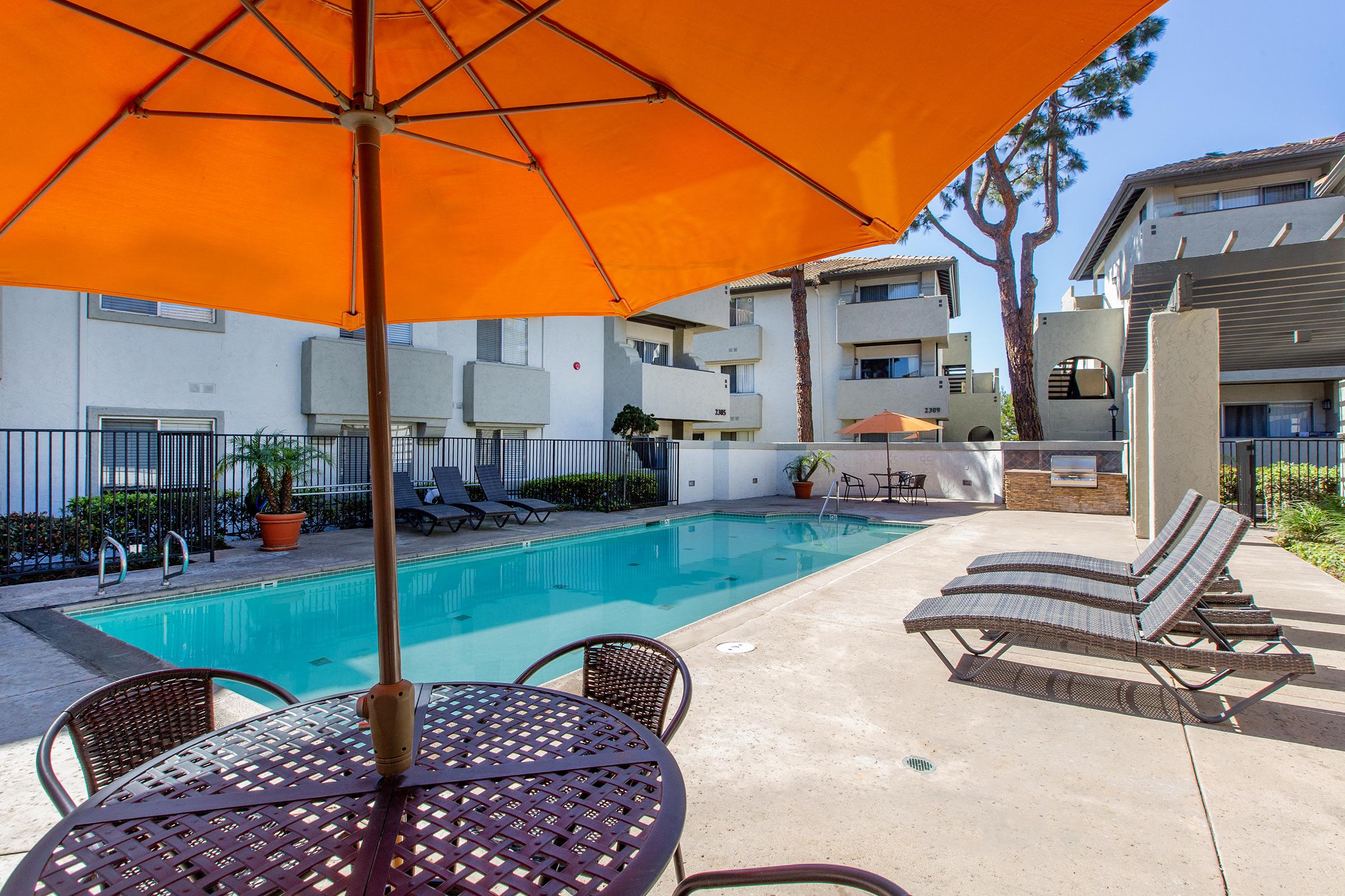 A sunny pool area featuring a clear blue swimming pool surrounded by lounge chairs and a table with an orange umbrella. Nearby are apartment buildings and lush greenery, creating a relaxing outdoor environment.