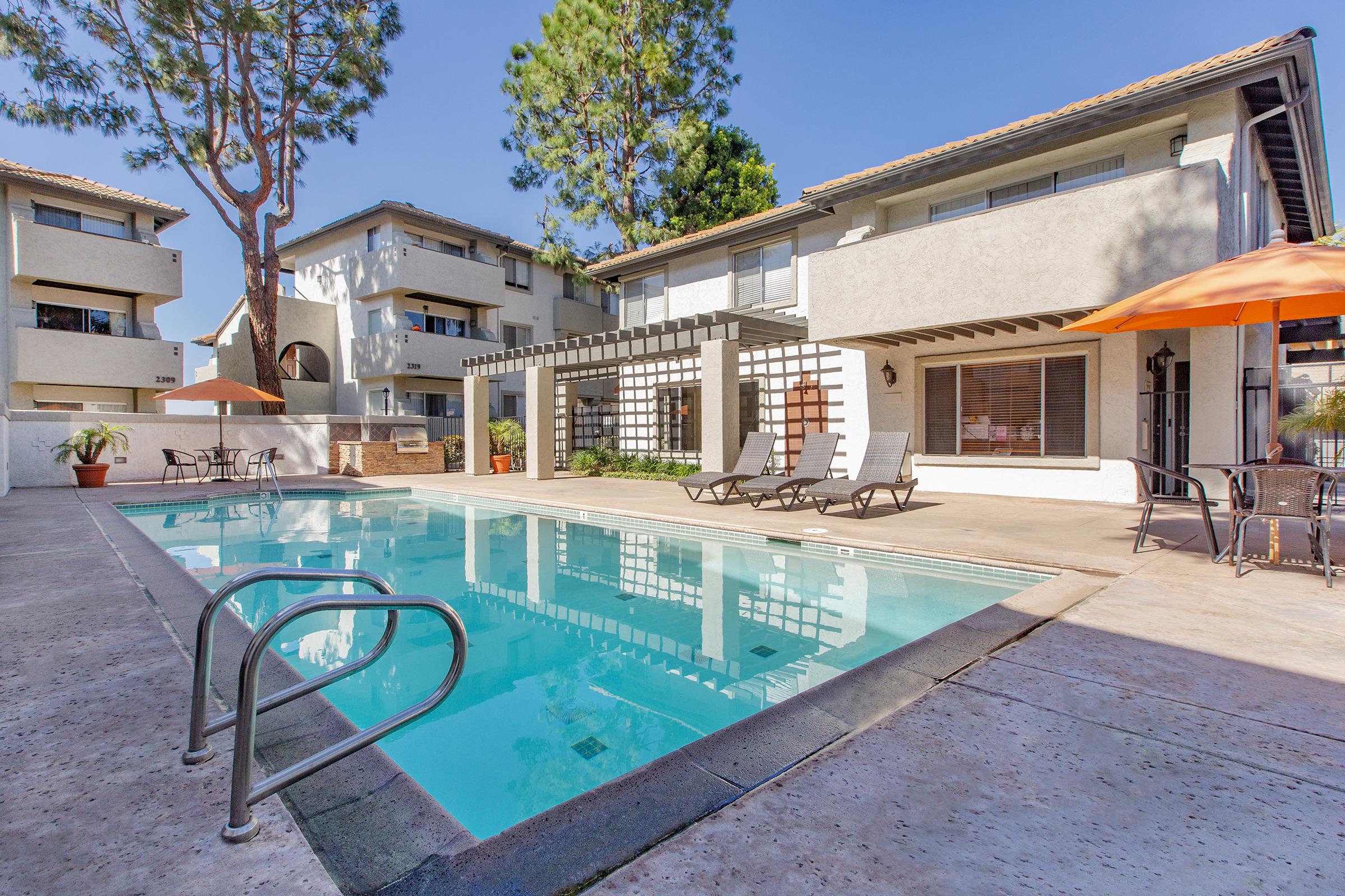 A bright and inviting apartment complex pool area featuring a clear blue pool surrounded by lounge chairs. There are shaded tables, tropical plants, and two-story buildings in the background under a clear blue sky, creating a relaxed outdoor atmosphere.