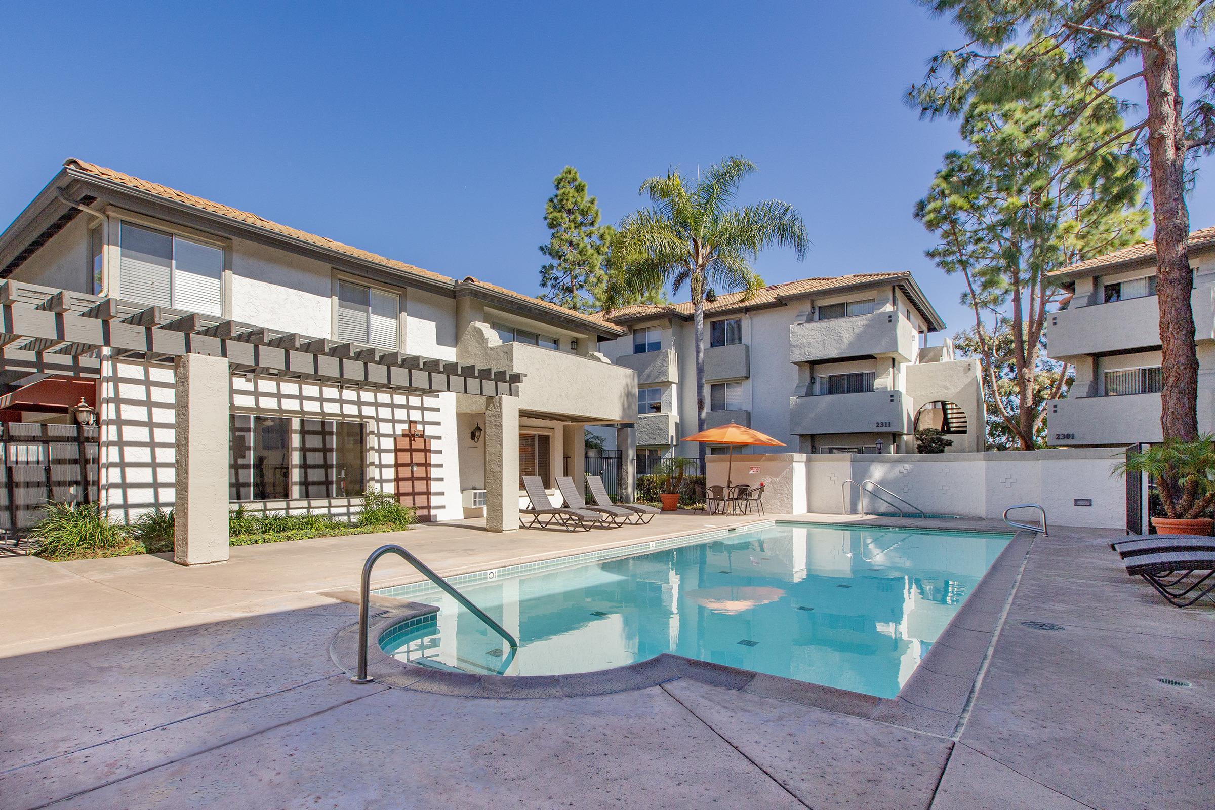 A sunny outdoor swimming pool area featuring a clear blue pool, lounge chairs, and an orange umbrella. Surrounding the pool are landscaped plants and a backdrop of modern buildings with balconies and a pergola structure. Towering palm trees add to the tropical ambiance.