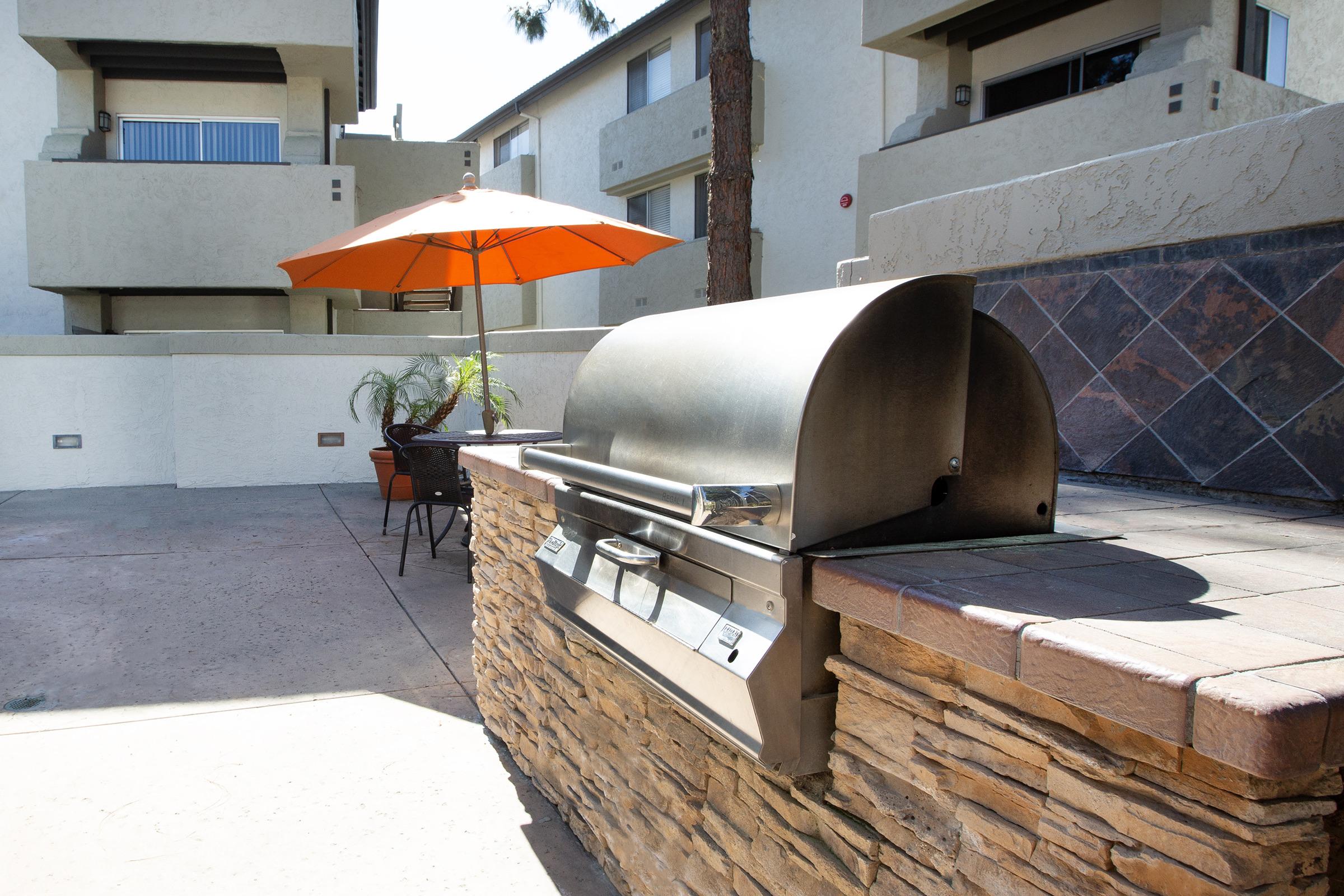 A stainless steel barbecue grill is set on a stone countertop in an outdoor area. An orange umbrella provides shade for a nearby table and chairs, surrounded by a landscaped environment. The backdrop showcases apartment buildings and palm trees.