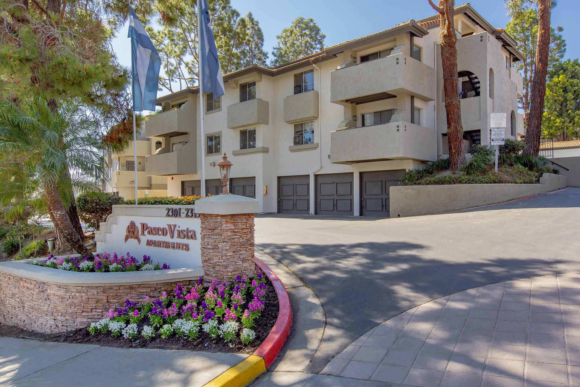 A residential building named "Paseo Vista" is shown, featuring a multi-story structure with balconies. Beautiful landscaping with flowers and plants surrounds the entrance, and there are several flags displayed. The driveway leads to garage doors at the base of the building, set amidst trees and greenery.