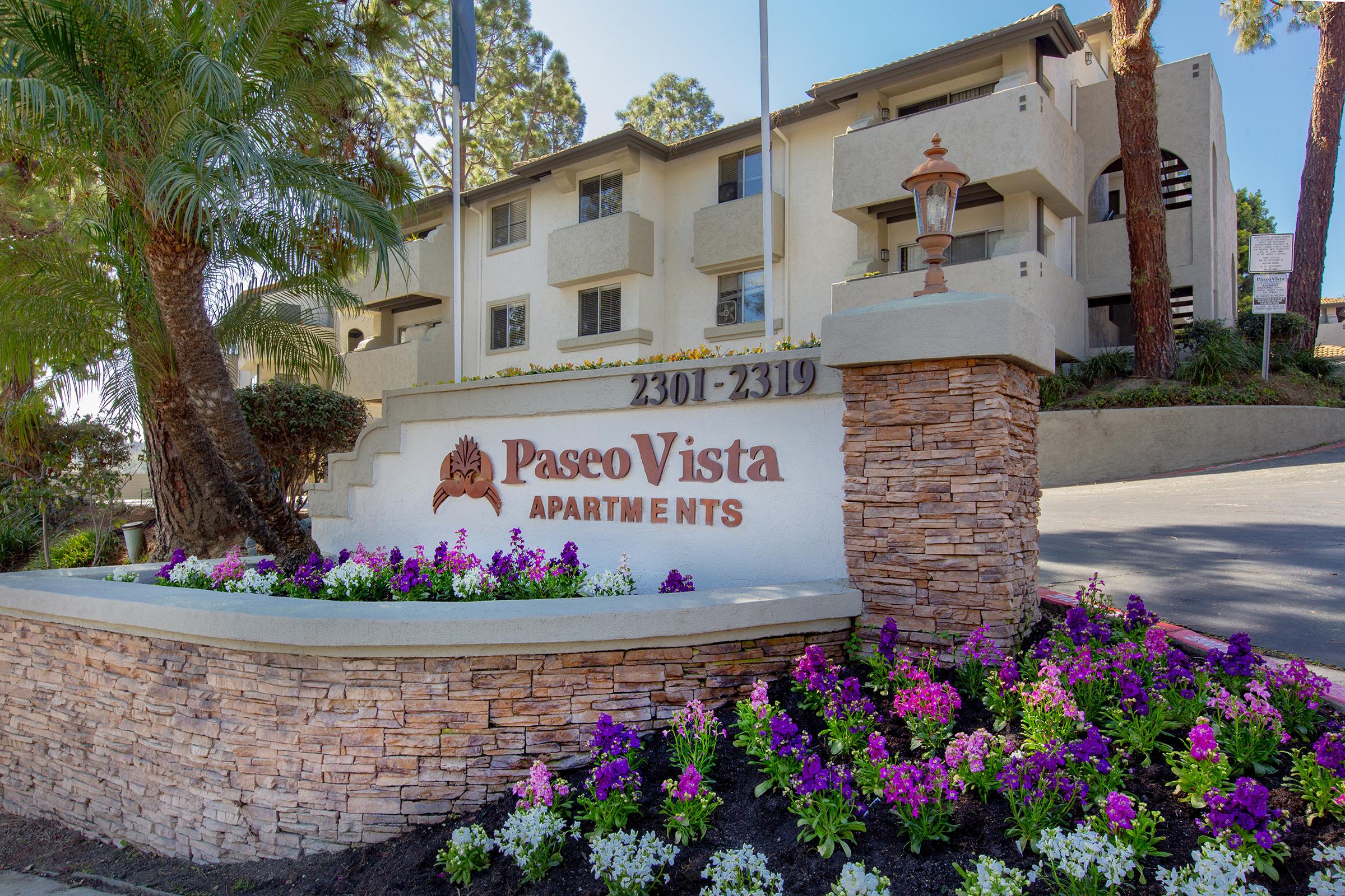 Sign for Pasco Vista Apartments with the address 3301-2319, surrounded by colorful flowers and greenery, set against a modern building in a sunny outdoor setting.
