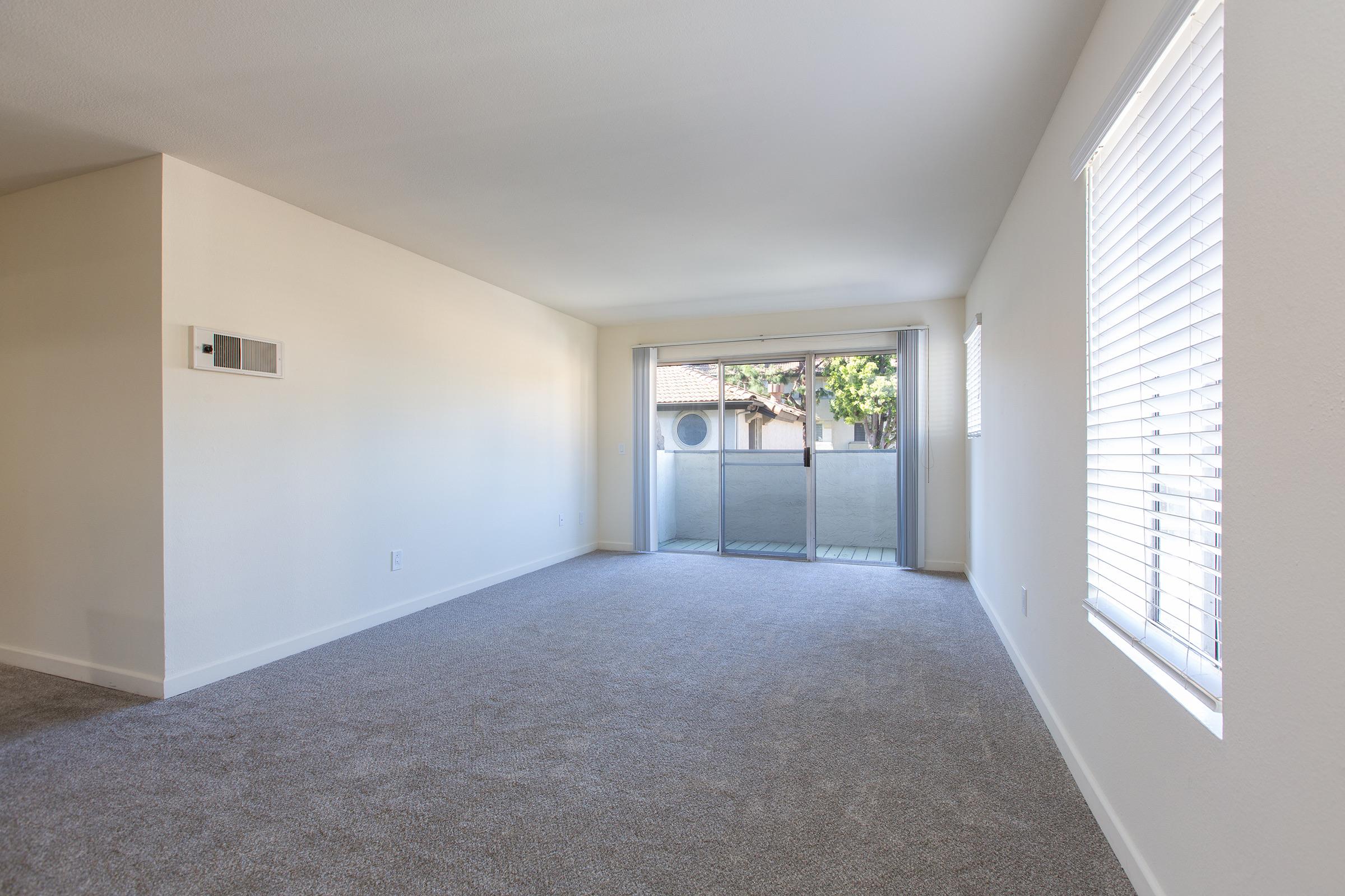 A spacious, empty living room featuring light-colored walls and carpet. A large sliding glass door leads to a balcony with views of greenery outside. Natural light streams in through two windows with blinds, creating a bright and airy atmosphere. The room has a minimalist design, ready for potential furnishings.