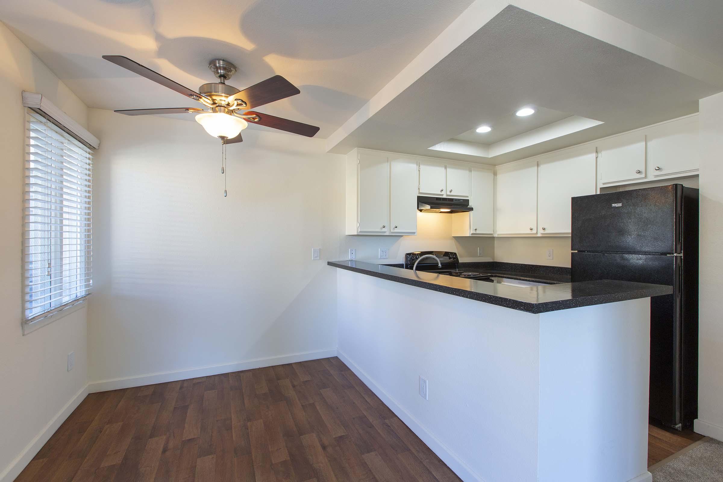 A modern kitchen with a ceiling fan, featuring white cabinets, black countertops, and appliances. There is a window with blinds allowing natural light, and hardwood-style flooring complements the space. The kitchen includes a black refrigerator and a stove, set in an open layout.