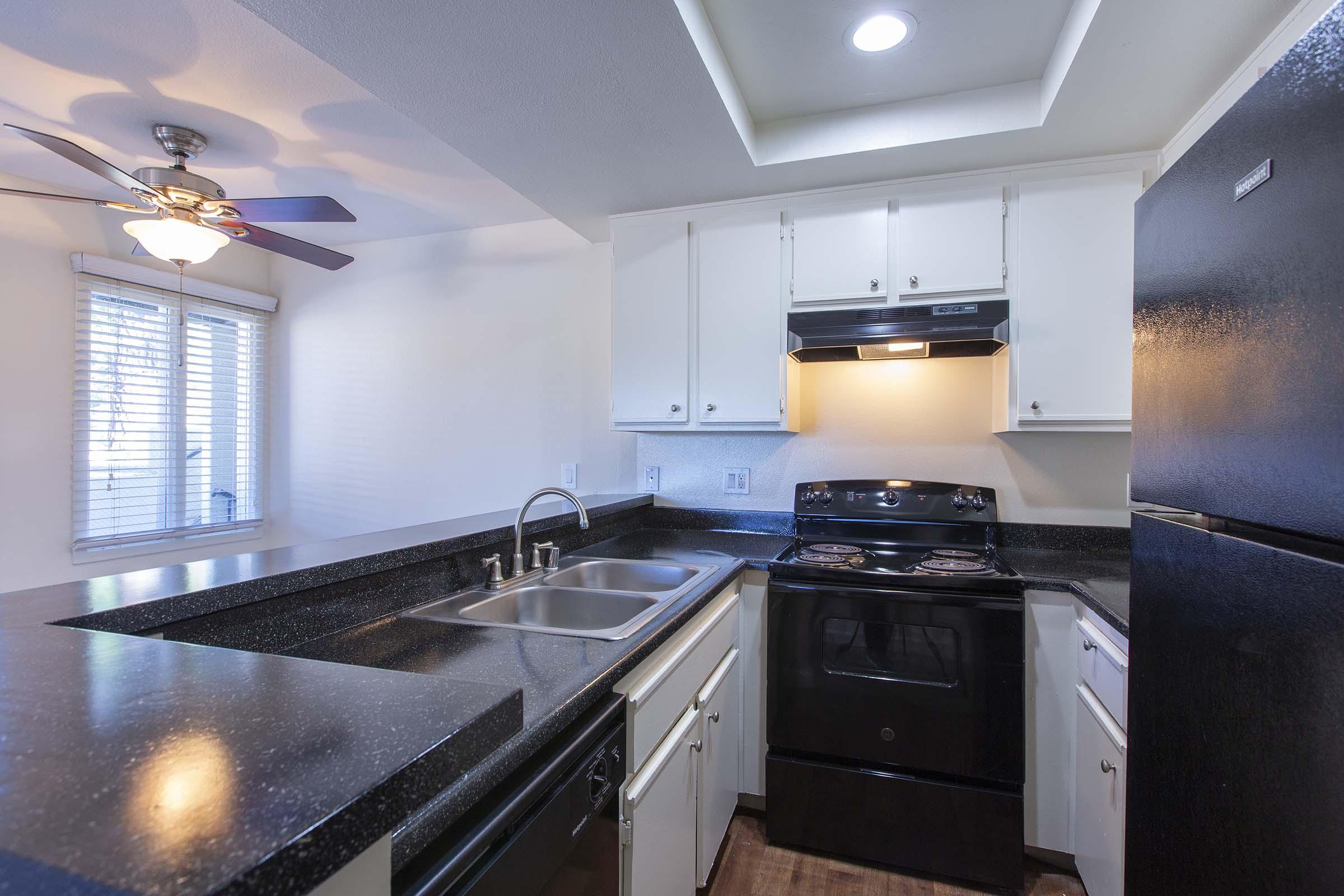 A modern kitchen featuring black appliances, including a stove and refrigerator, set against white cabinetry. The countertops are dark, and there’s a double sink. A ceiling fan and a window with blinds provide lighting and ventilation. The overall design is sleek and contemporary.