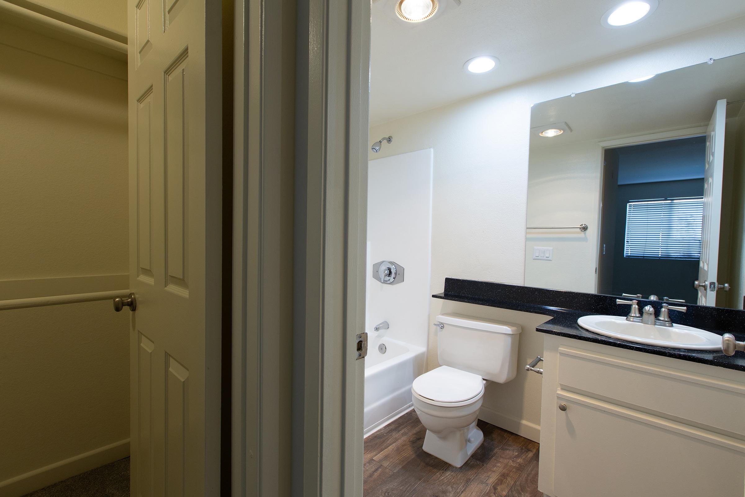 A clean and modern bathroom featuring a bathtub with shower, a toilet, and a sink with a large mirror. On the left, a doorway leads to a small closet. The floor has a warm wooden appearance, and natural light filters in through a nearby window. The walls are painted in neutral colors.