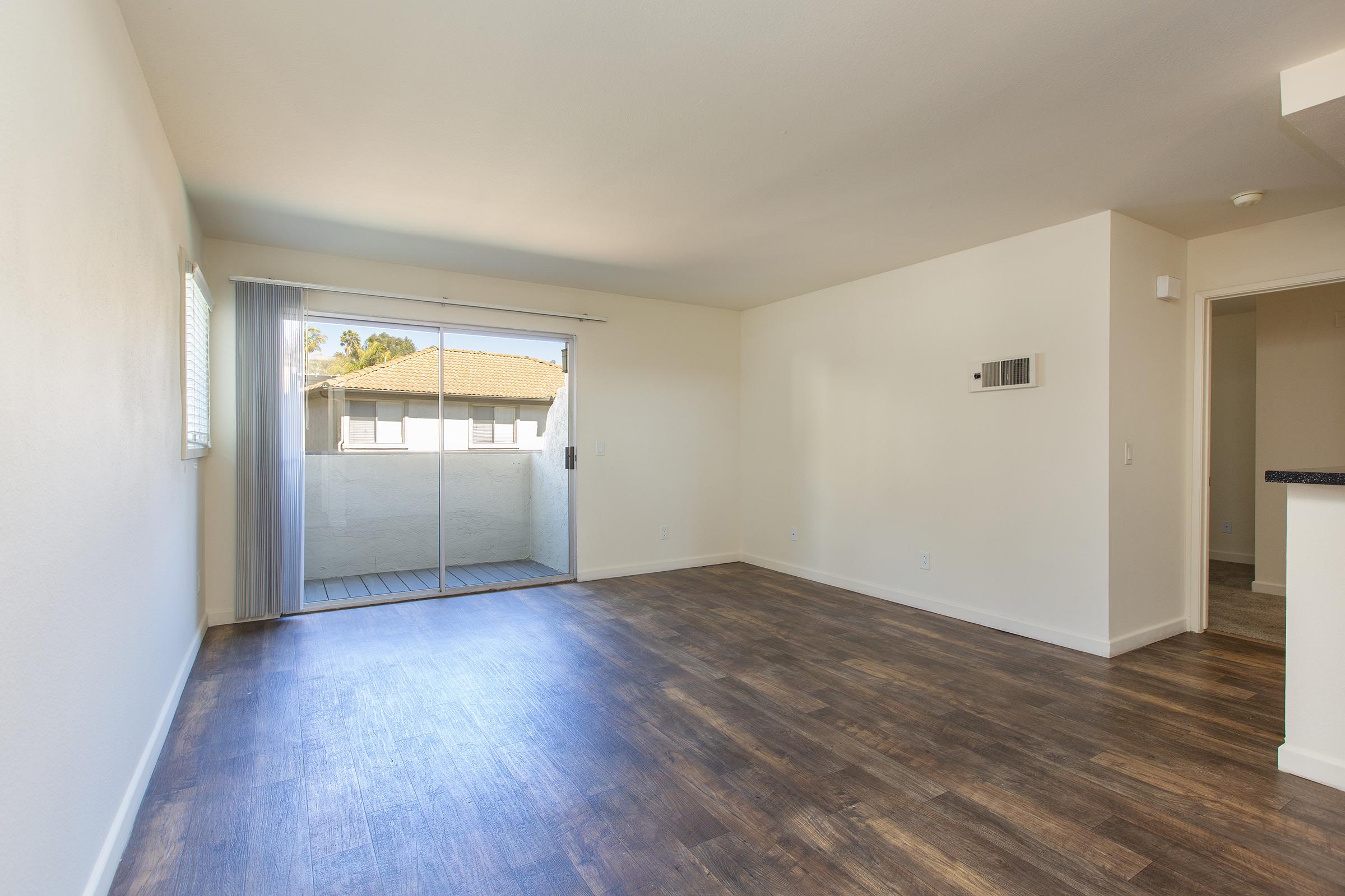 Empty interior of an apartment featuring light-colored walls, vinyl flooring, and a large sliding glass door leading to a small balcony. Sunlight enters through the window, illuminating the space. A kitchen area is visible in the background, with minimal furnishings.