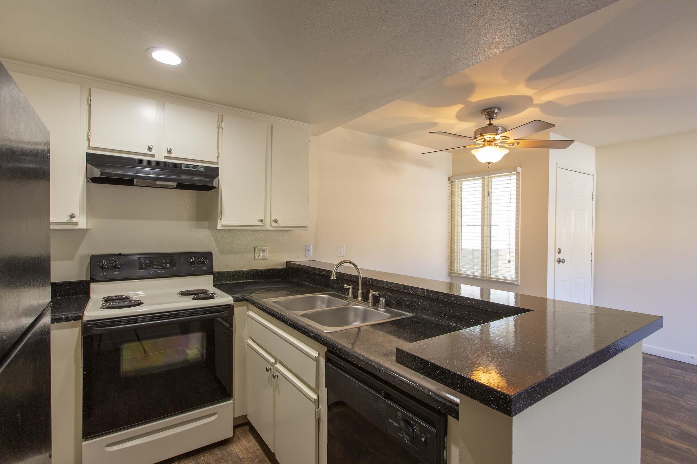 A modern kitchen featuring white cabinets, black countertops, and stainless steel appliances. The space includes a stove, oven, and sink, with a ceiling fan and windows providing natural light. The open layout leads to a living area with a doorway visible in the background.