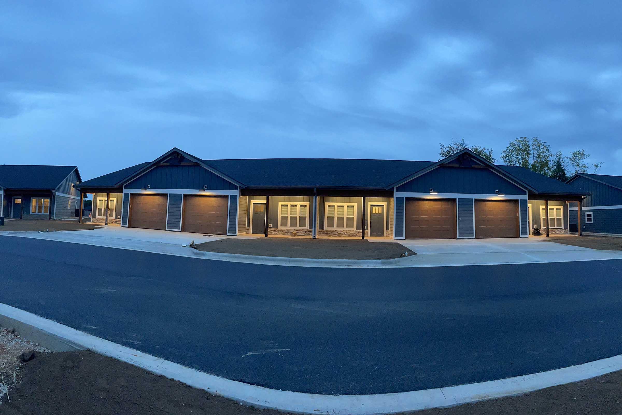 A modern building complex featuring multiple garage doors and a dark exterior with warm interior lighting. The sky is overcast, creating a moody atmosphere. The structure is situated on a paved circular driveway, surrounded by grassy areas, suggesting a suburban setting.