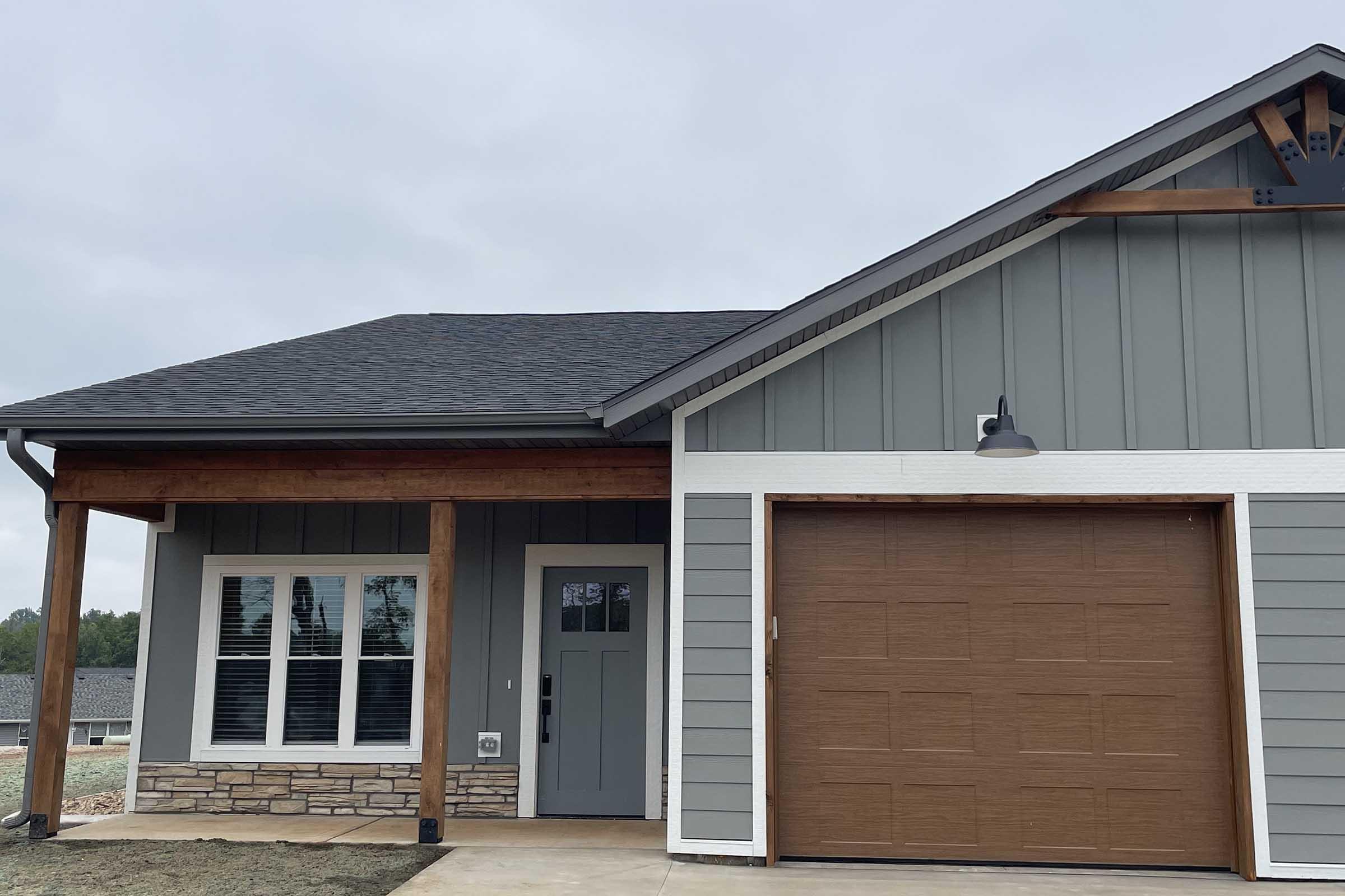 A modern house exterior featuring gray siding, a stone entryway, and a brown garage door. The front porch is supported by wooden beams, and there are large windows with white trim. The sky is cloudy, and the landscaping is minimal.