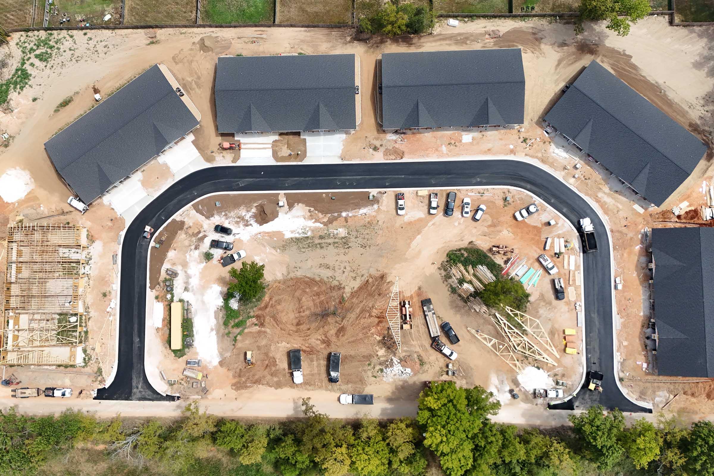 Aerial view of a developing residential area featuring several new buildings with dark roofs arranged along a curved road. Construction materials and machinery are visible on-site, along with parked vehicles. Surrounding greenery indicates the area is under development amidst natural surroundings.