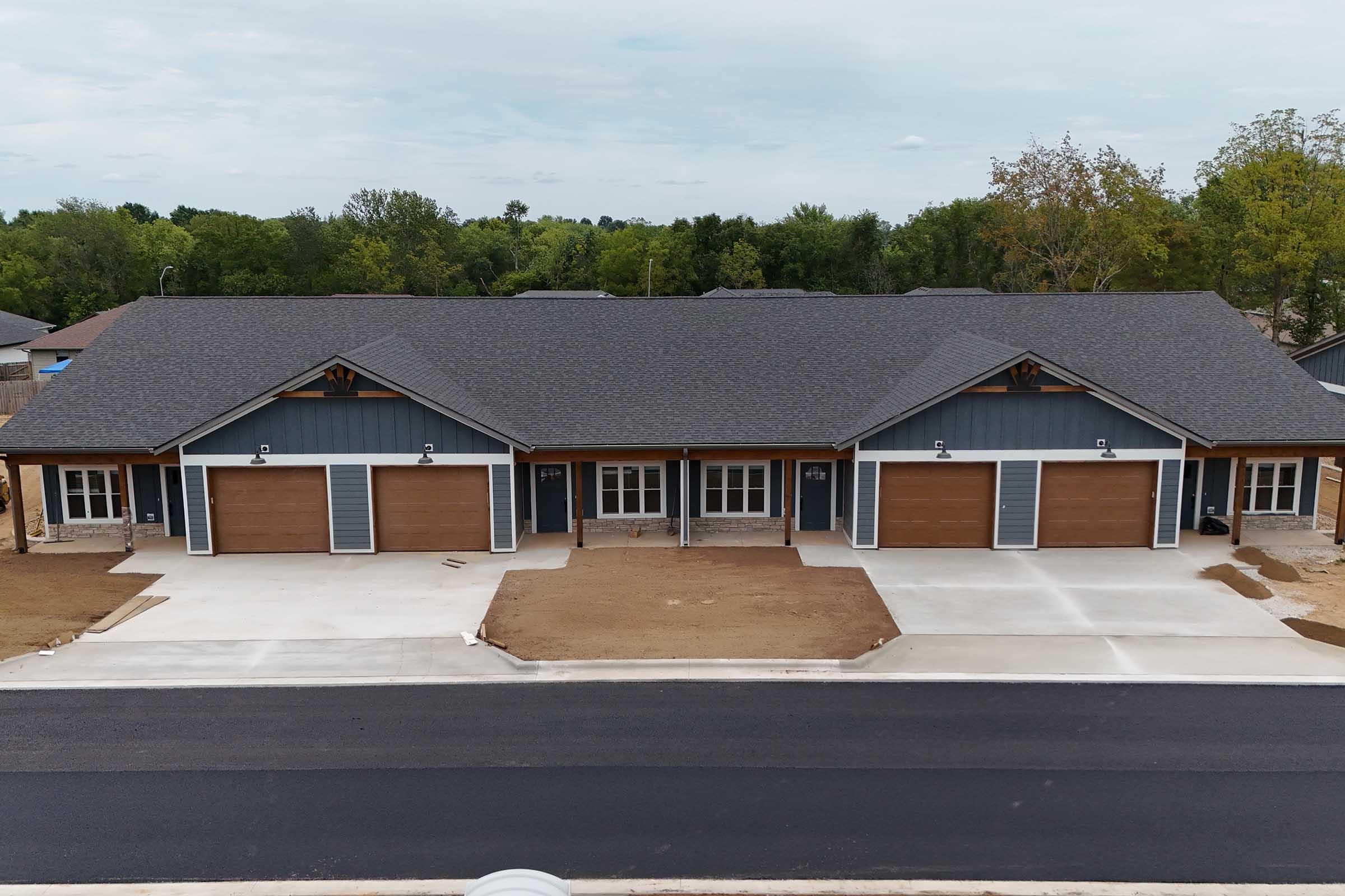 A modern single-story building with a symmetrical design, featuring four garage doors and a central entrance. The structure has a mix of gray siding and wooden accents, surrounded by a freshly landscaped area and asphalt road. Trees and greenery are visible in the background under a cloudy sky.