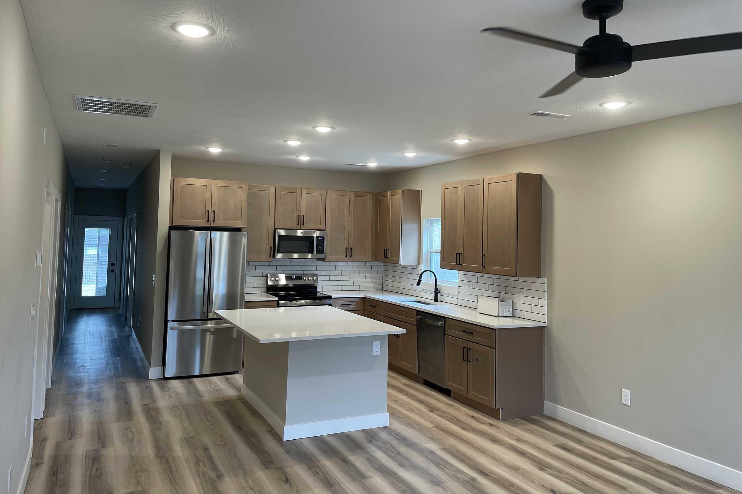 Modern kitchen featuring wooden cabinetry, stainless steel appliances, a large island with a white countertop, and a tiled backsplash. The space is well-lit with recessed lighting, and has an open layout leading to a hallway. The floor is finished in light-colored wood laminate.