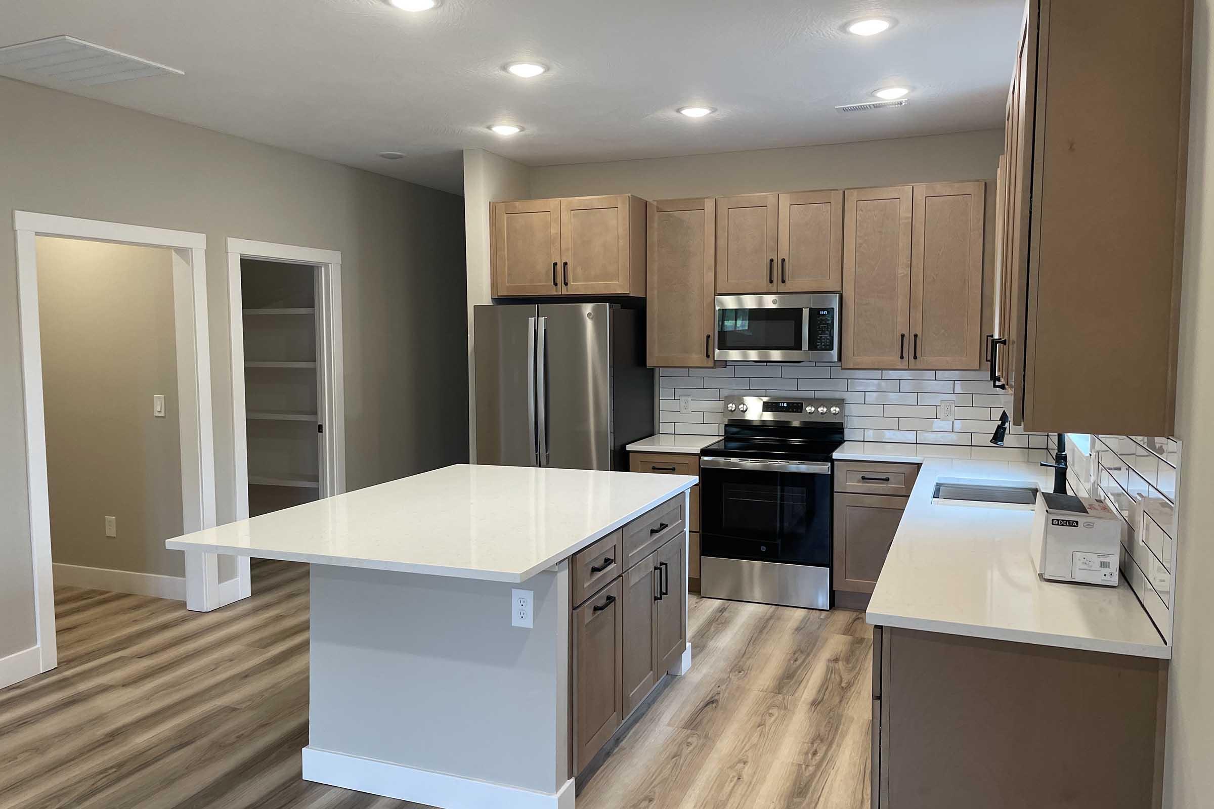 Modern kitchen with light brown cabinetry, stainless steel appliances, and a large island with white countertops. Features include a gas stove, microwave, and a spacious pantry visible through an open door. The flooring is light wood, and the walls are painted a soft gray, creating a warm and inviting atmosphere.