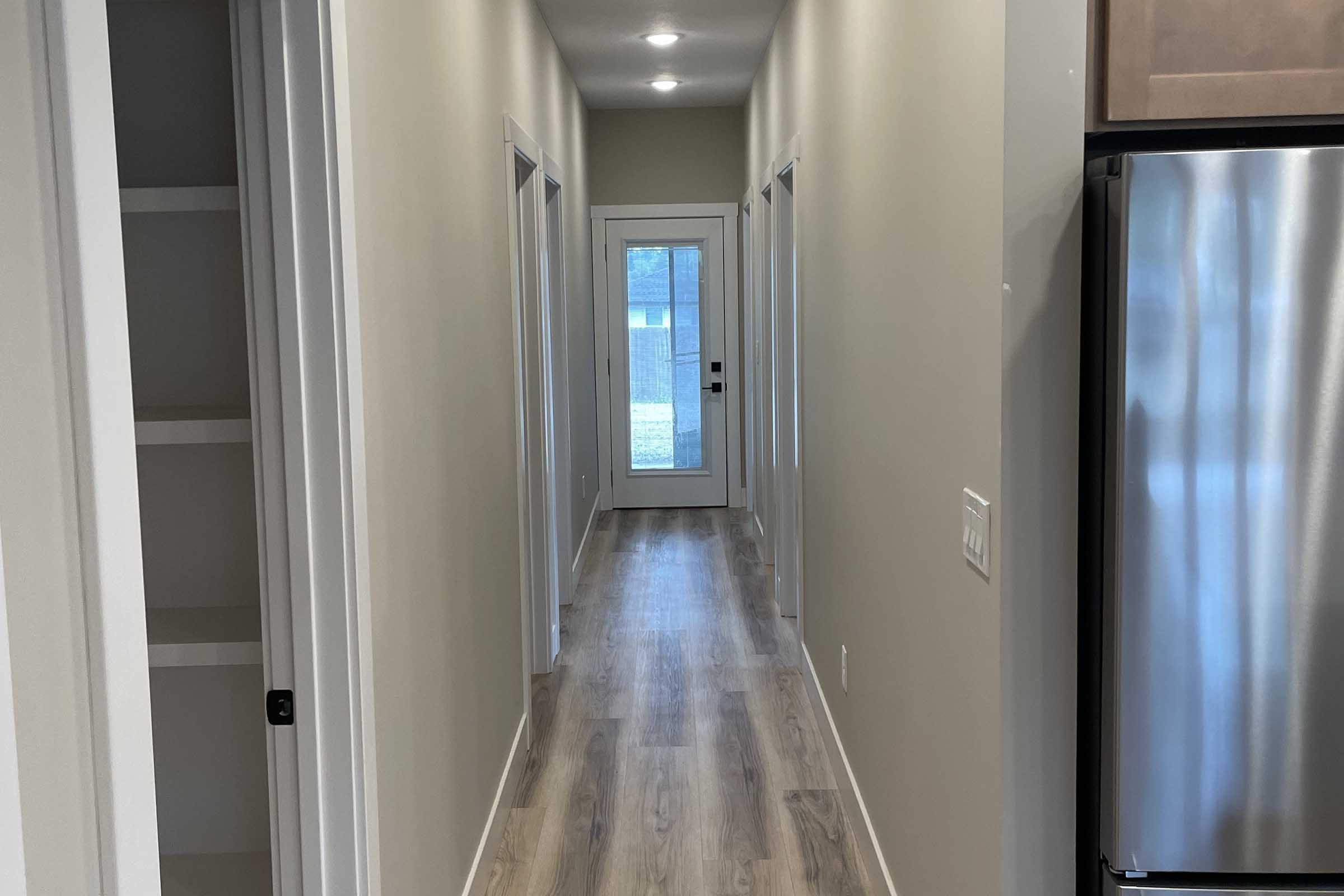 A well-lit, modern hallway featuring light-colored walls and a light wooden floor. On one side, there is a door leading outside, and on the other, a closet space is visible. The hallway is lined with doors leading to other rooms, creating a spacious and inviting atmosphere.