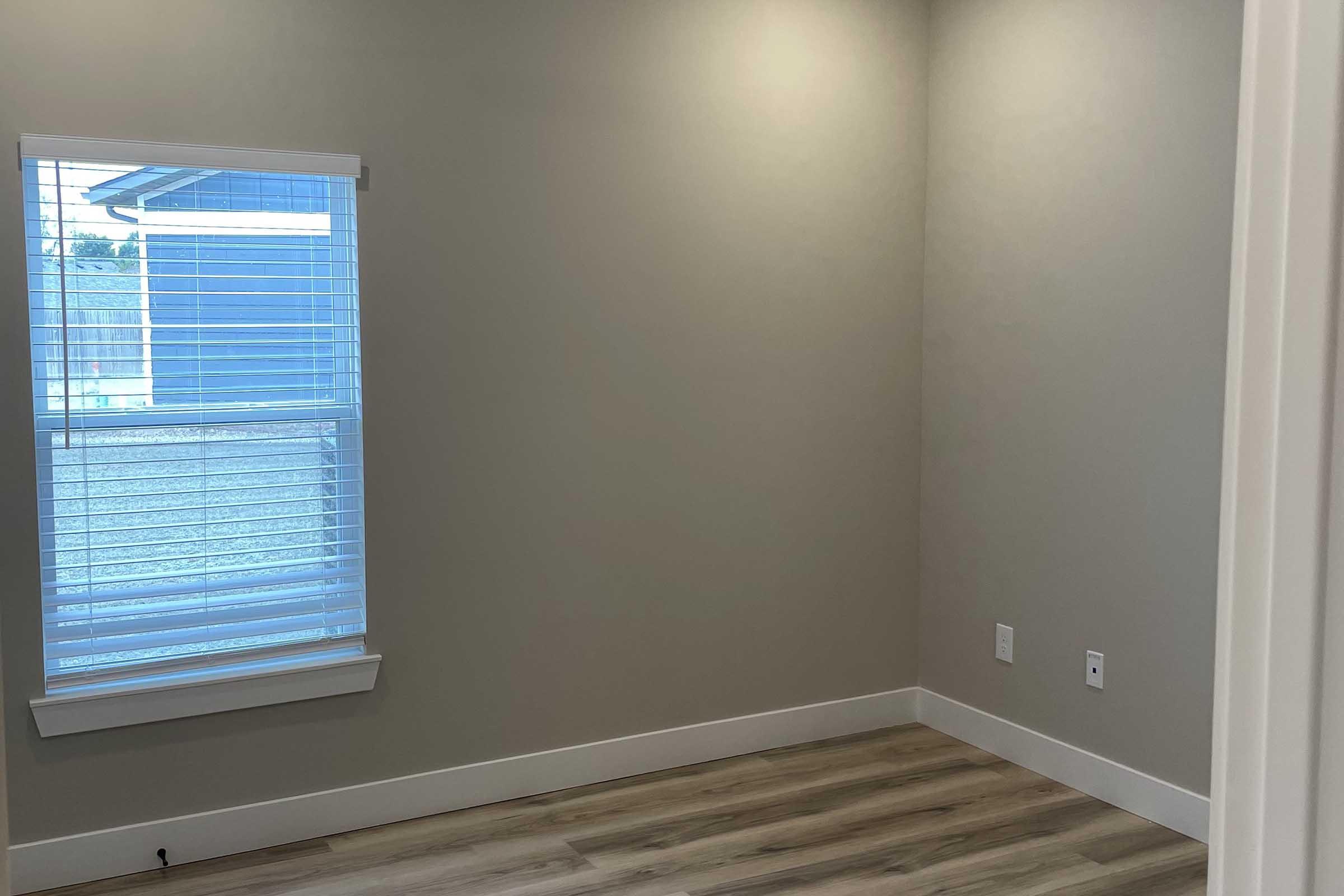 A vacant room with light gray walls and light hardwood flooring. It features a window with white blinds that lets in natural light, casting a subtle glow in the space. The room is unfurnished, creating an open and airy feel, ideal for personalization.