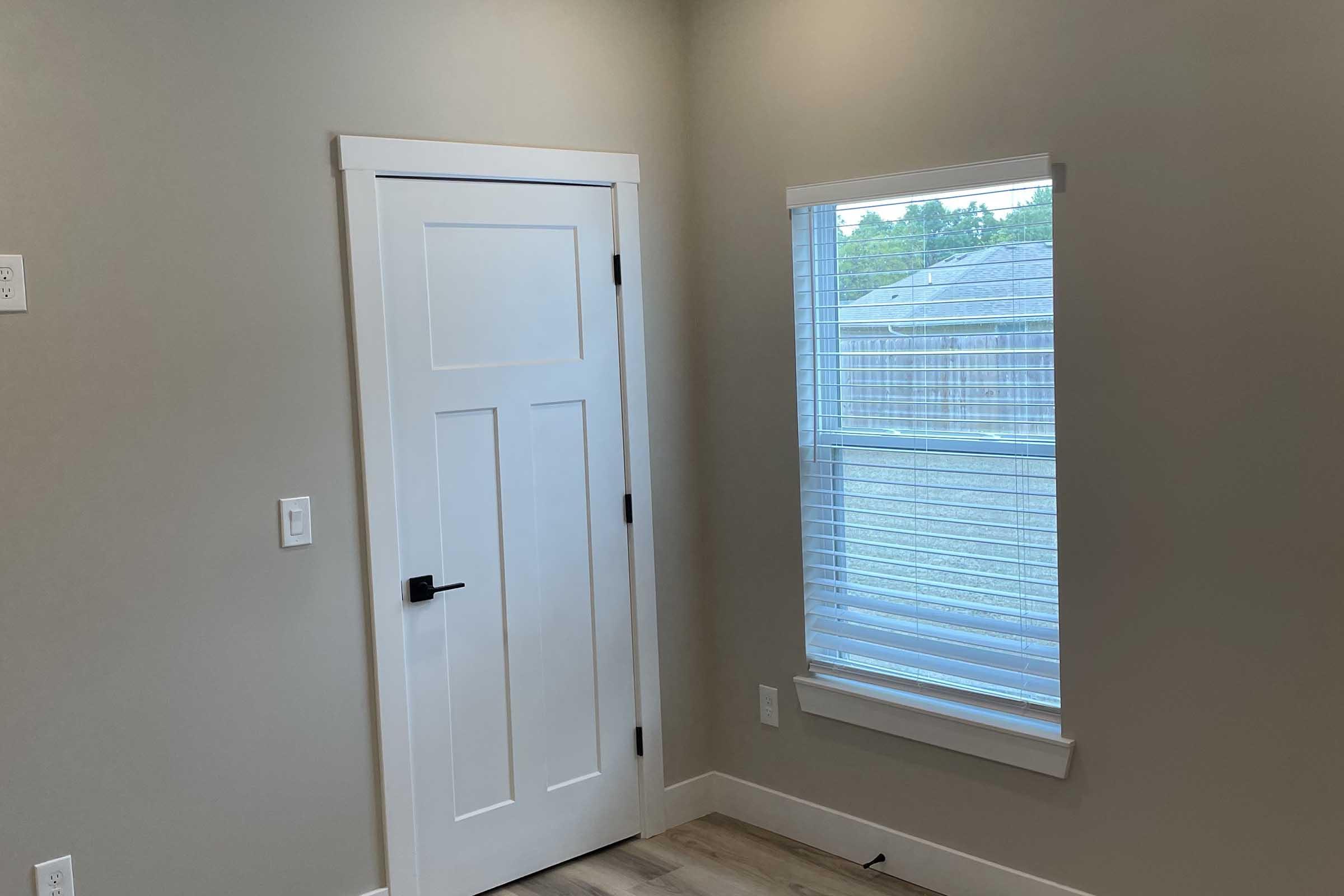 A well-lit interior corner of a room featuring a closed white door and a window with blinds. The walls are painted a light gray, and the floor is wooden. The window offers a view of the outdoors, with light streaming in, creating a bright atmosphere.