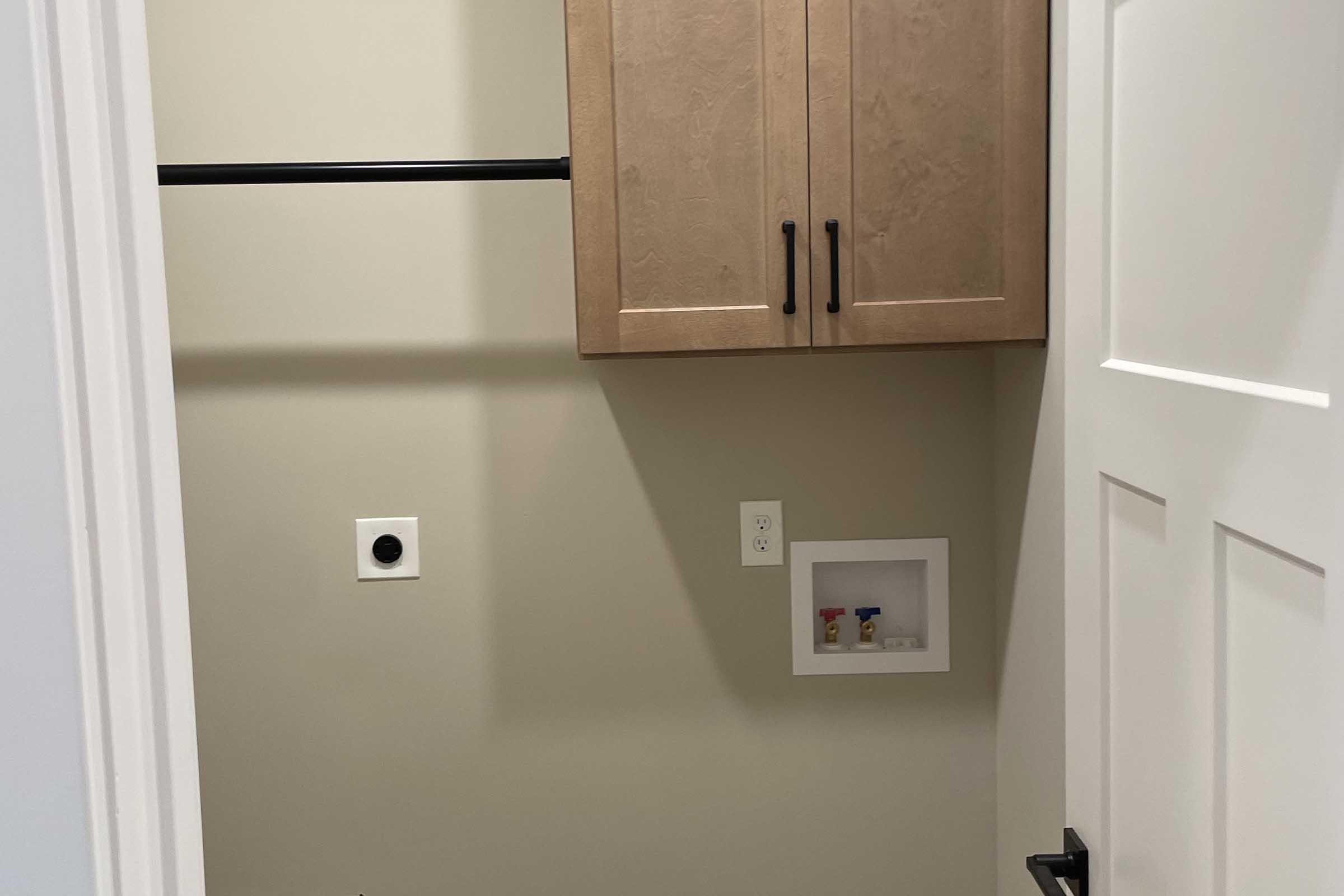 A small, empty room featuring a white door, beige walls, a wooden cabinet with two doors mounted above a closet rod, and a built-in shelf with two small items visible. An electrical outlet and a circular vent are also visible on the wall. The space appears clean and organized.