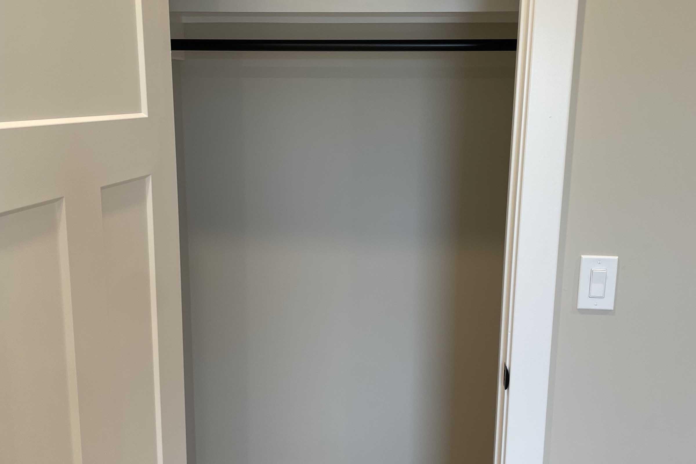 An empty closet with a single hanging rod, featuring a light gray interior and white doors. The right side shows a light switch on the wall.