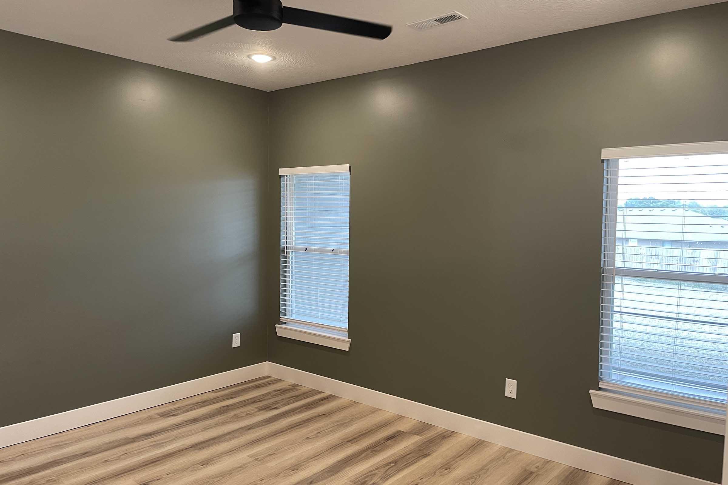 A spacious room with olive green walls, featuring two windows with horizontal blinds. The floor is made of light-colored wood, and there is a ceiling fan mounted in the center. Natural light enters the room, creating a bright and airy atmosphere.