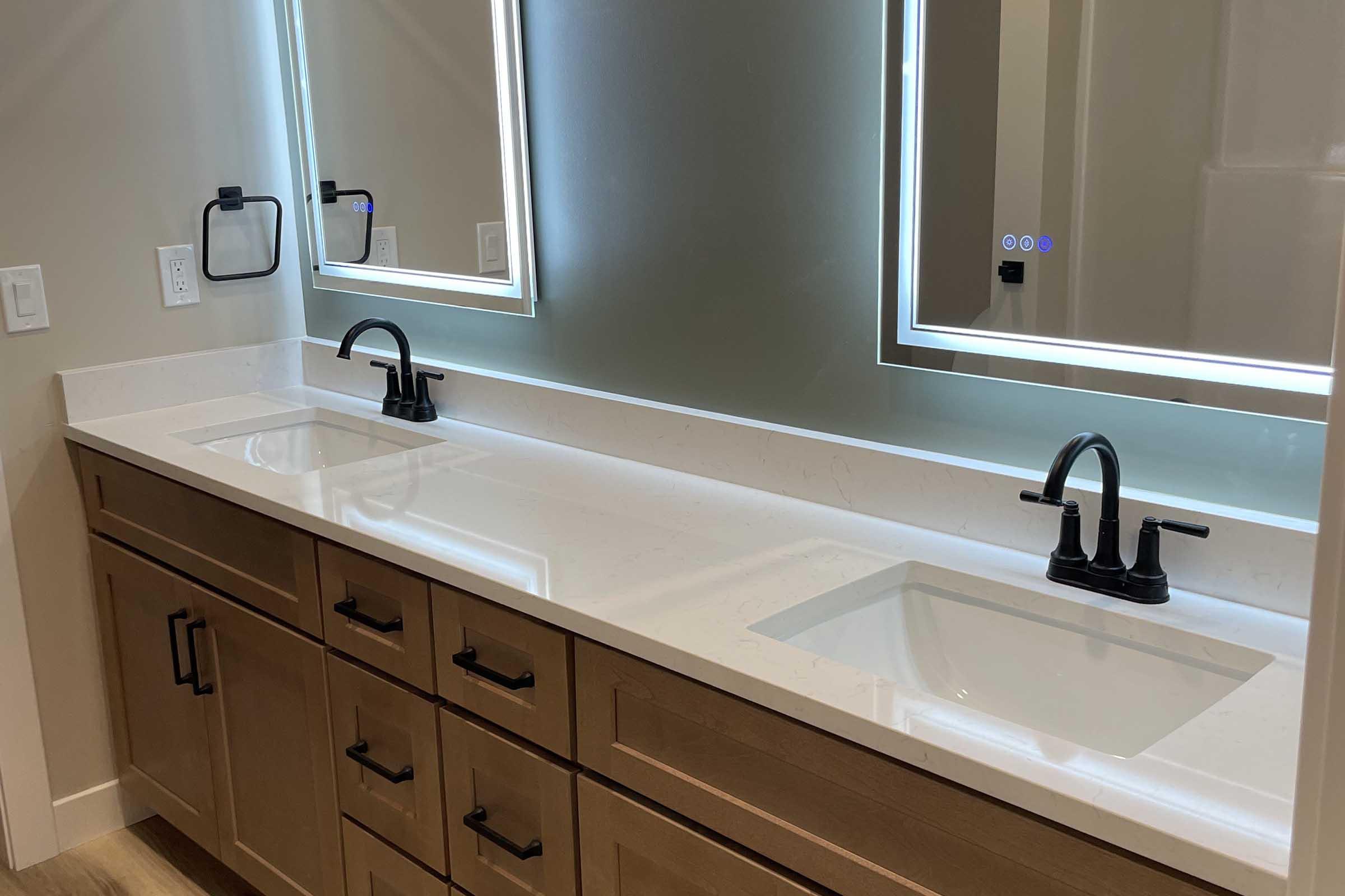 Modern bathroom featuring a double sink vanity with black faucets and mounted mirrors. The vanity has a light-colored countertop and wooden cabinetry. Soft lighting enhances the contemporary design, creating a clean and bright ambiance.
