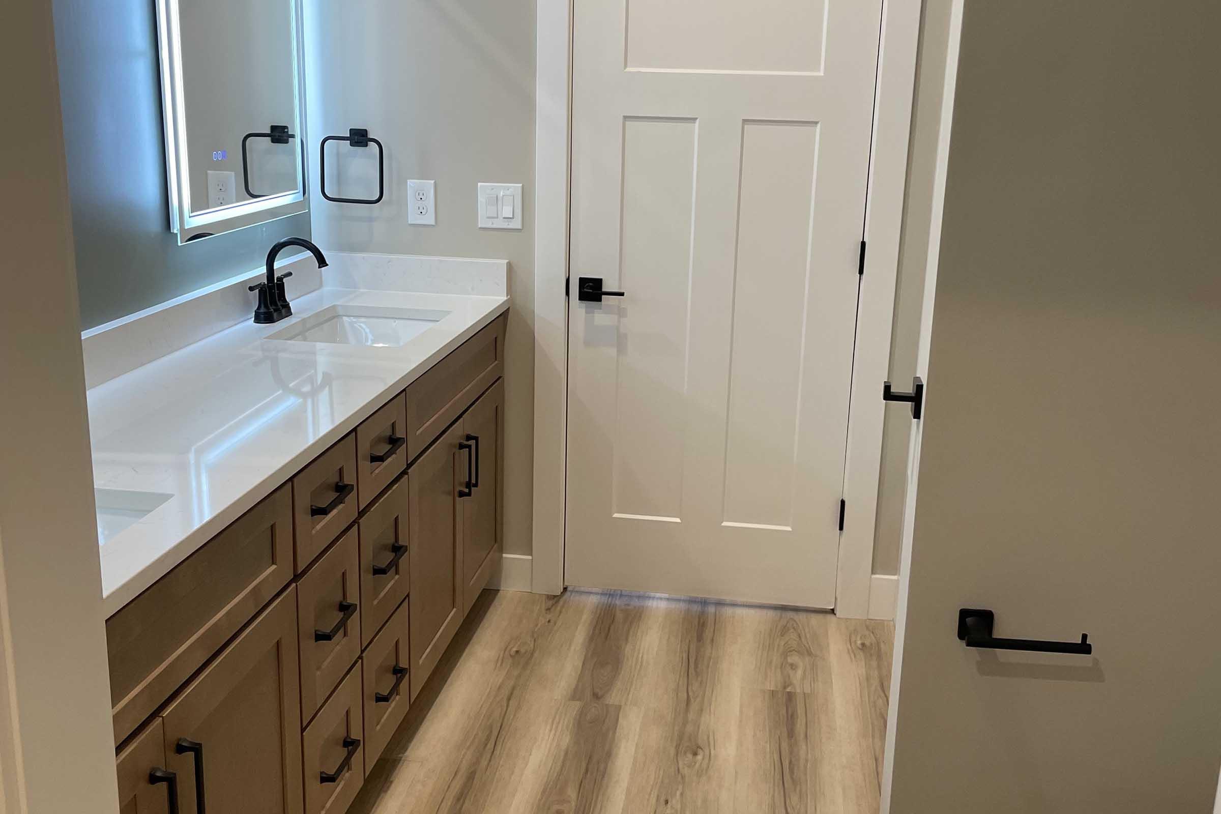 A modern bathroom interior featuring a double sink vanity with brown cabinetry, a white countertop, and a large mirror. The walls are painted a light color, and there is a closed door leading to another room. The floor consists of wooden planks, adding warmth to the space.