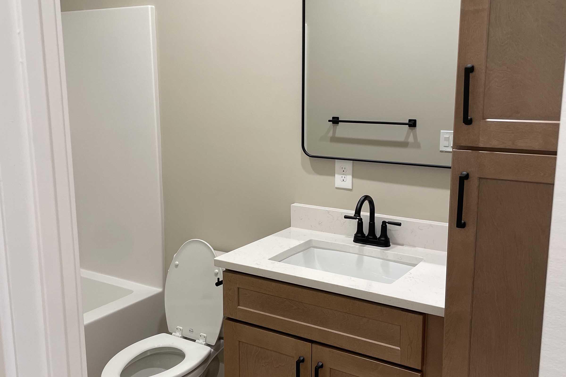 A modern bathroom featuring a white bathtub, a compact vanity with a square sink, and a black faucet. There is a large mirror above the sink and a black towel rack on the wall. The cabinetry is light wood, and the walls are a soft neutral color.
