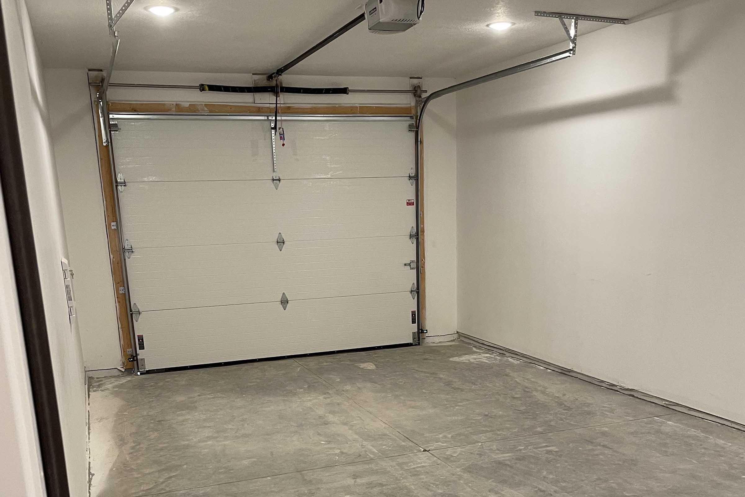A spacious, empty garage with a closed white garage door. The walls are unadorned and painted in neutral colors. The floor appears smooth and gray, and the ceiling features overhead lighting. The overall space is clean and uncluttered, ready for storage or vehicle parking.
