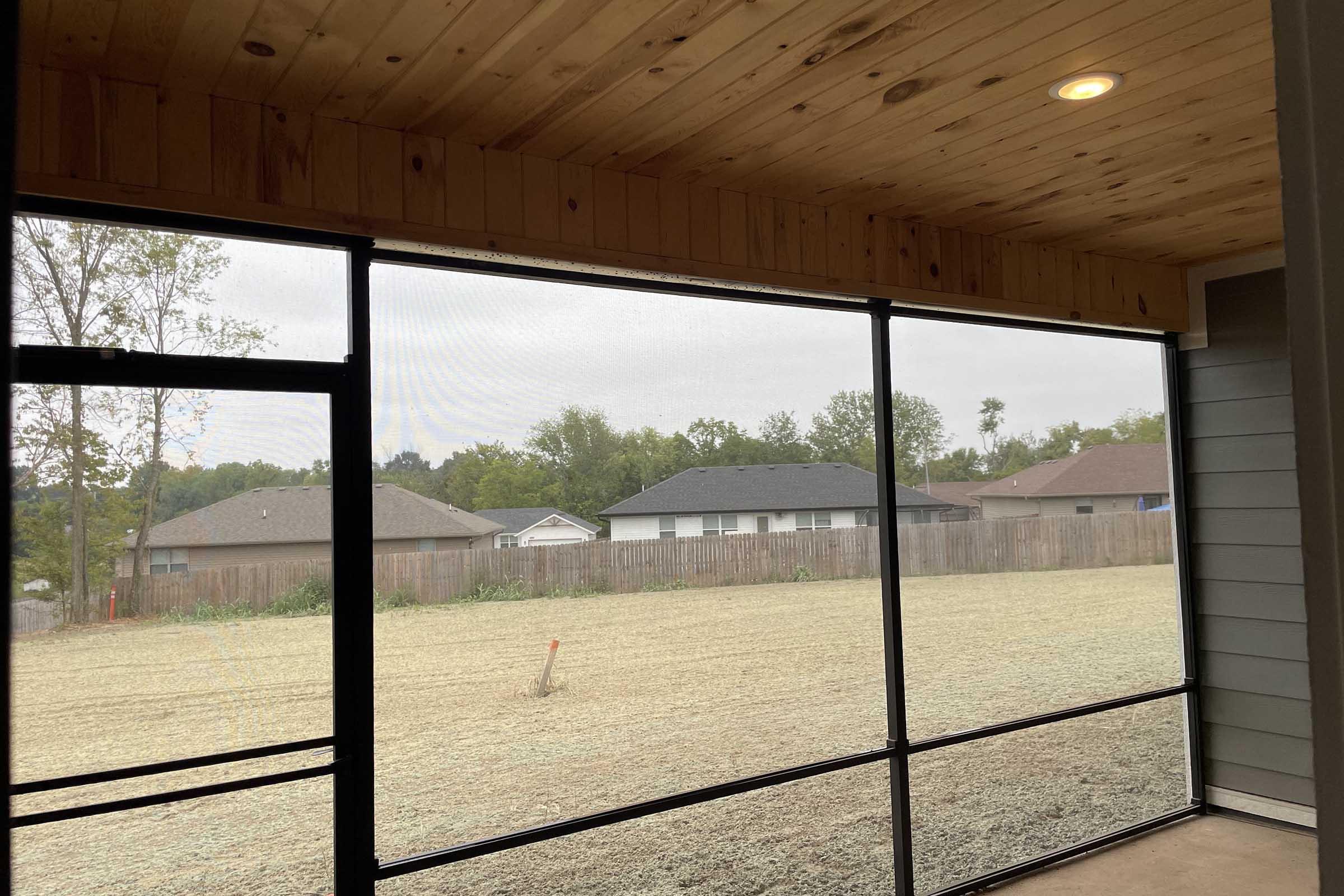 Interior view of a screened porch with wooden ceiling. Looking out through the screens, there is a grassy field and several houses in the distance, surrounded by trees. The sky is overcast, creating a calm and muted atmosphere.
