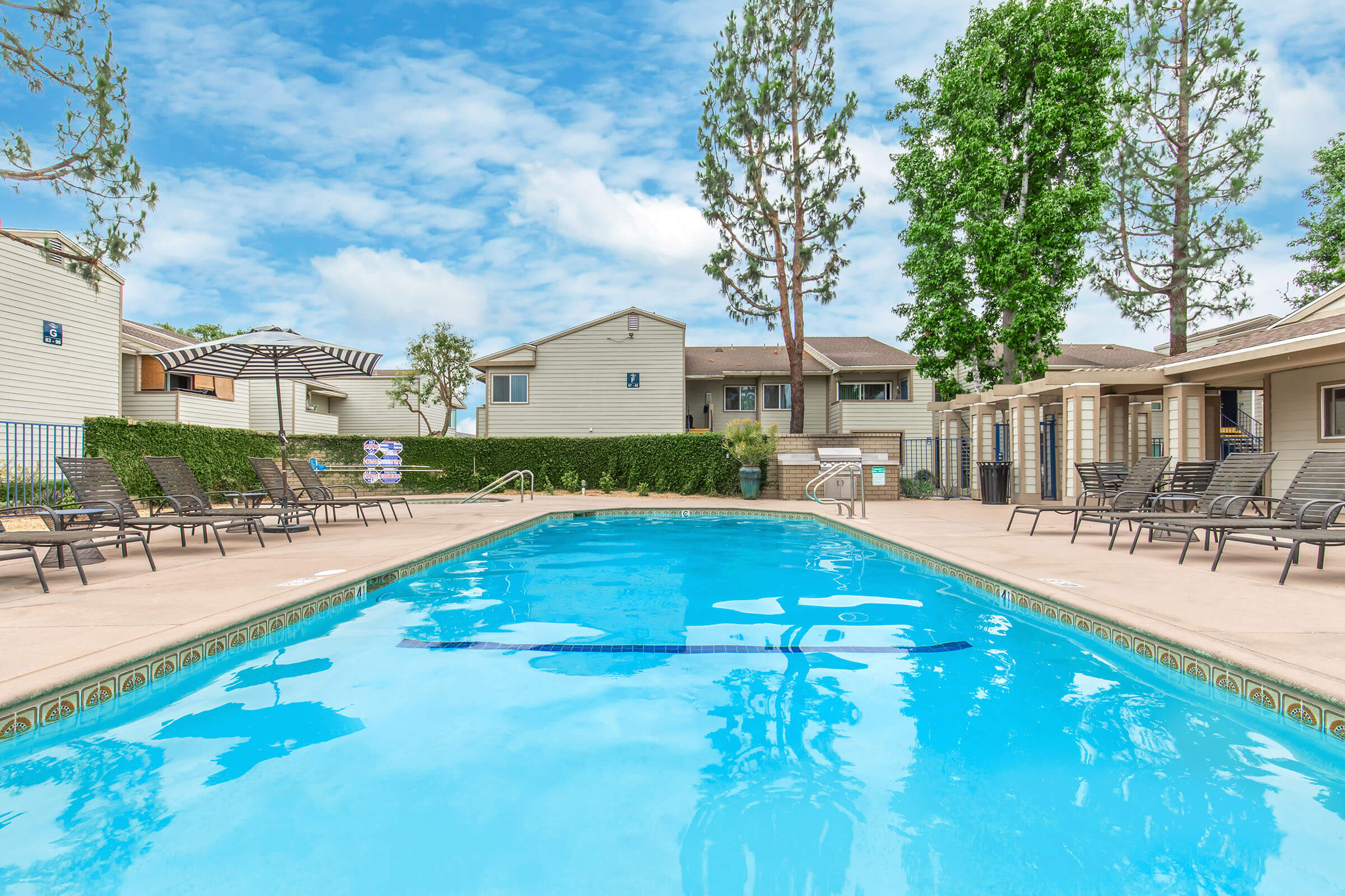 A clear swimming pool surrounded by lounge chairs and umbrellas, with residential buildings and tall trees in the background under a partly cloudy sky. The scene conveys a relaxing outdoor atmosphere.