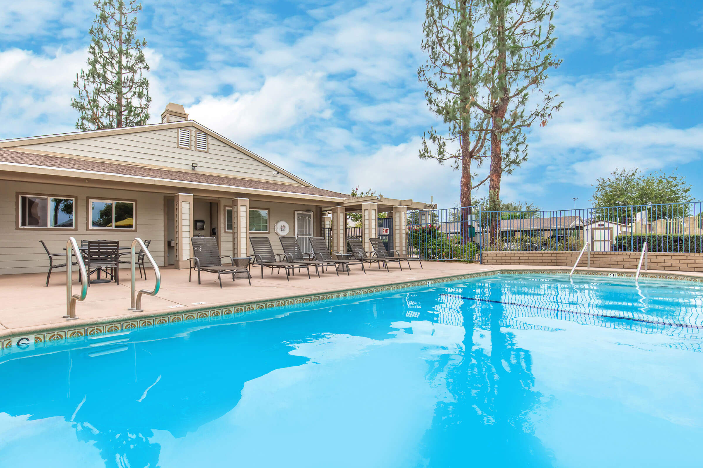 A clear blue swimming pool with lounge chairs around it, situated next to a modern building. In the background, tall trees are visible against a partly cloudy sky, creating a serene outdoor atmosphere. The area is well-maintained, suggesting a residential or recreational setting.