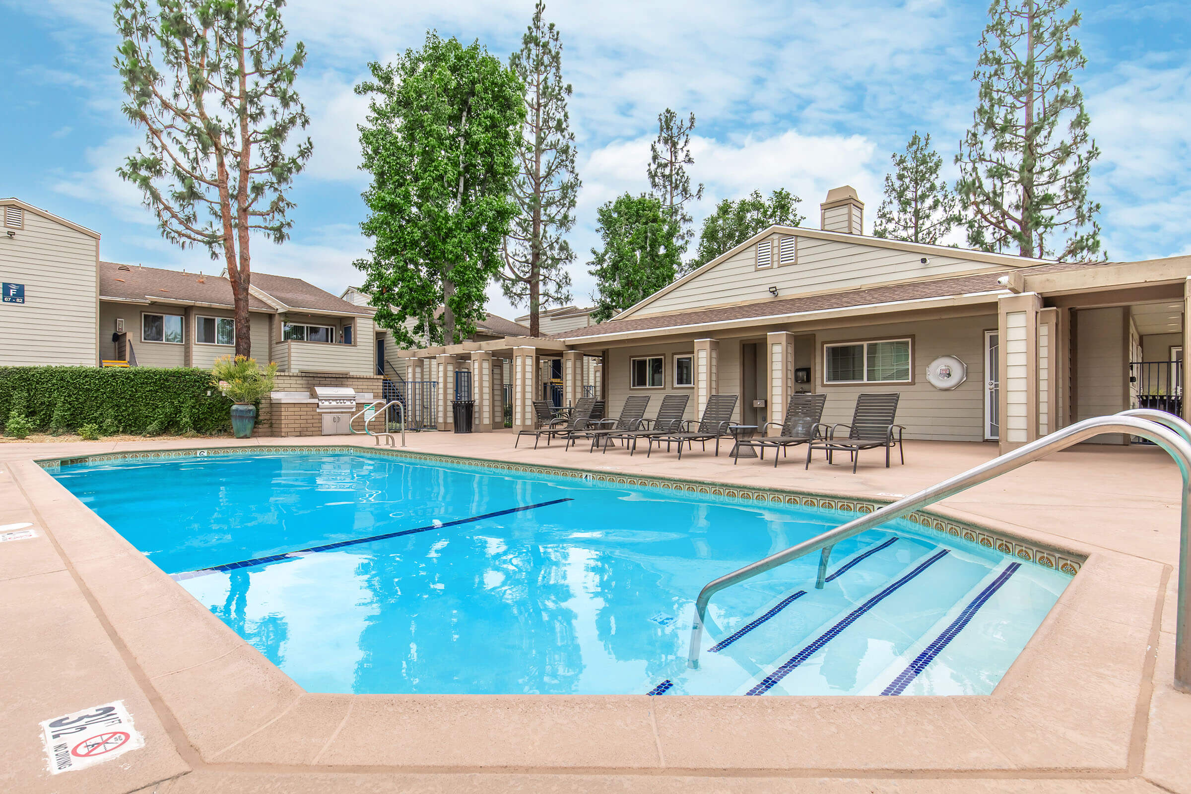 A clear blue swimming pool surrounded by lounge chairs, with trees and buildings in the background. The pool area is well-maintained, featuring a handrail leading into the water. There's a shaded area with a building that includes windows, offering a relaxing atmosphere.
