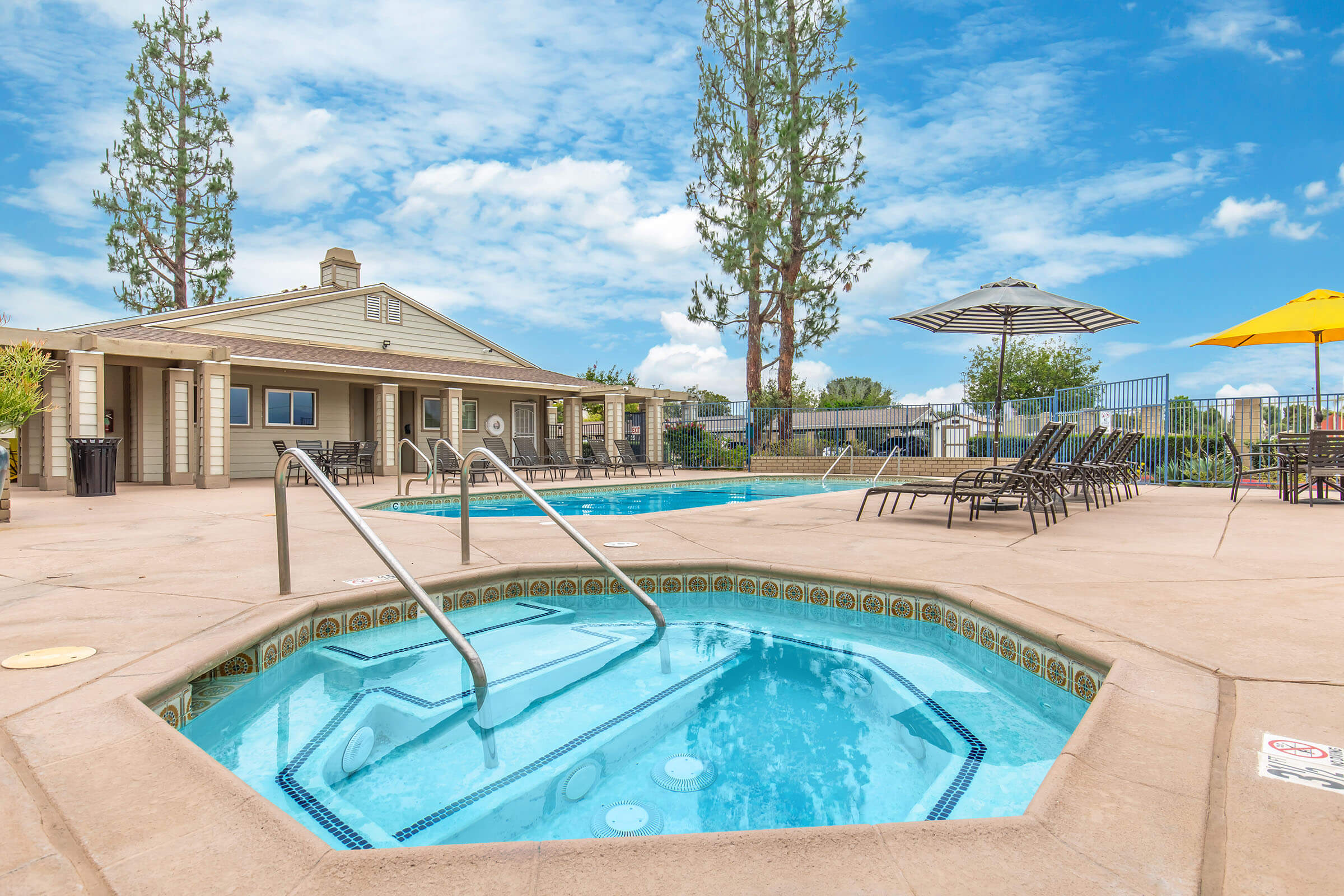 A scenic view of a swimming pool area featuring a hot tub in the foreground, surrounded by lounge chairs and umbrellas. In the background, a clubhouse is visible, framed by tall trees under a blue sky with fluffy clouds. The setting suggests a relaxing outdoor leisure space.