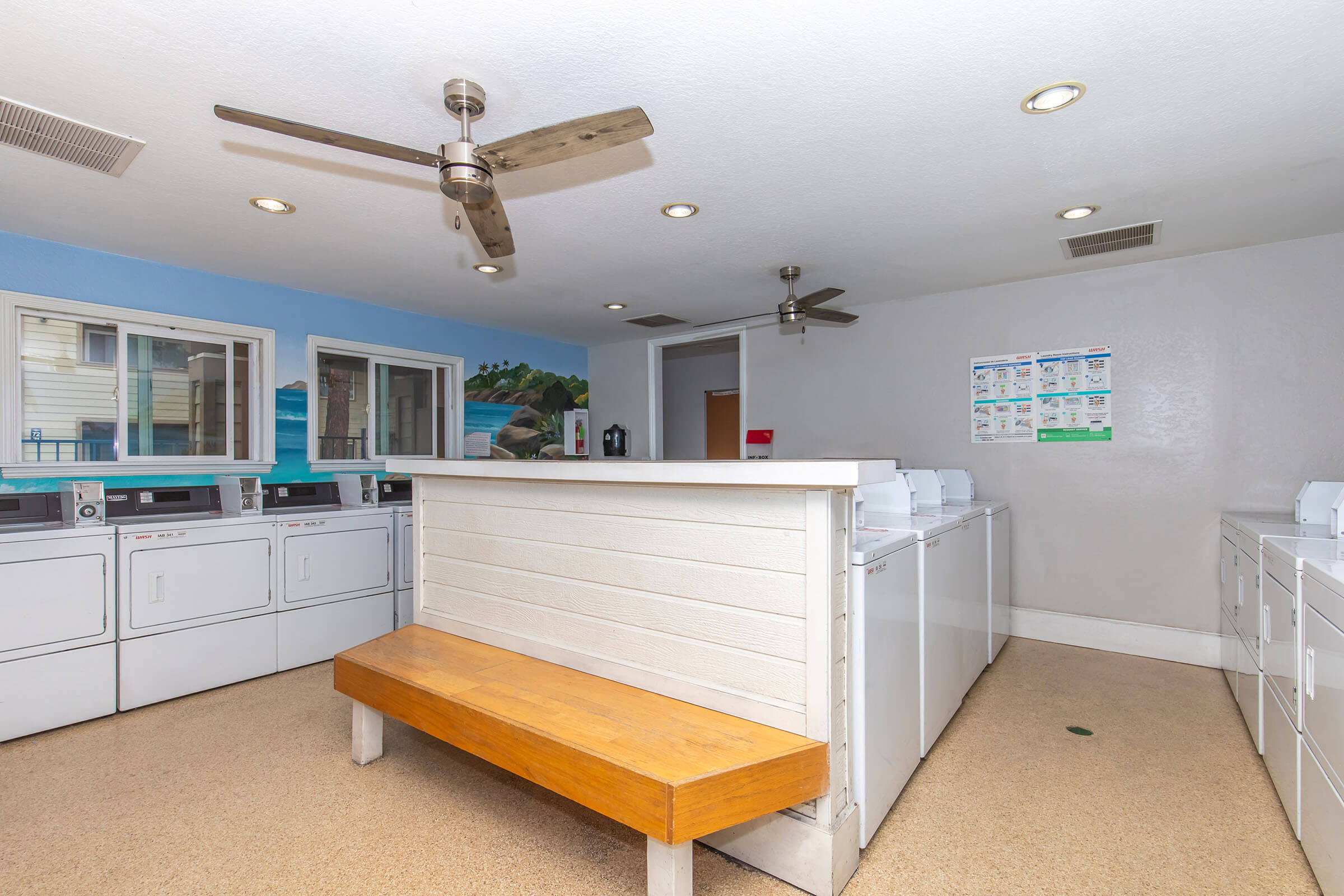 A clean and well-lit laundry room featuring multiple washing machines and dryers lined up against the wall. There's a wooden bench in the center, ceiling fans above, and large windows that provide natural light, with a mural of a beach scene on one wall.