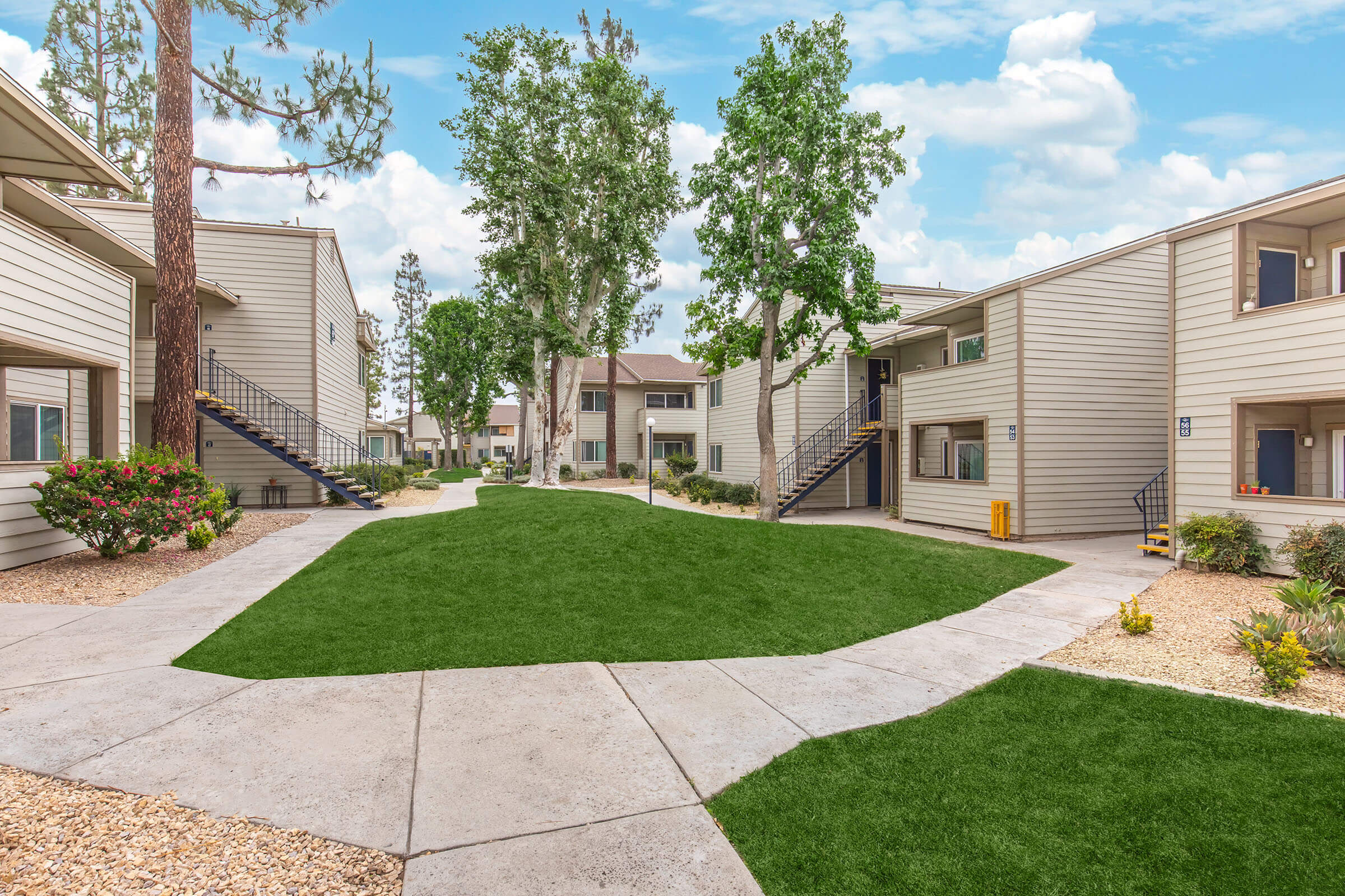 A landscaped courtyard featuring several two-story apartment buildings surrounded by green grass and shrubs. There are concrete pathways leading through the area, with trees providing shade in the background and a clear blue sky above. The setting appears well-maintained and inviting.