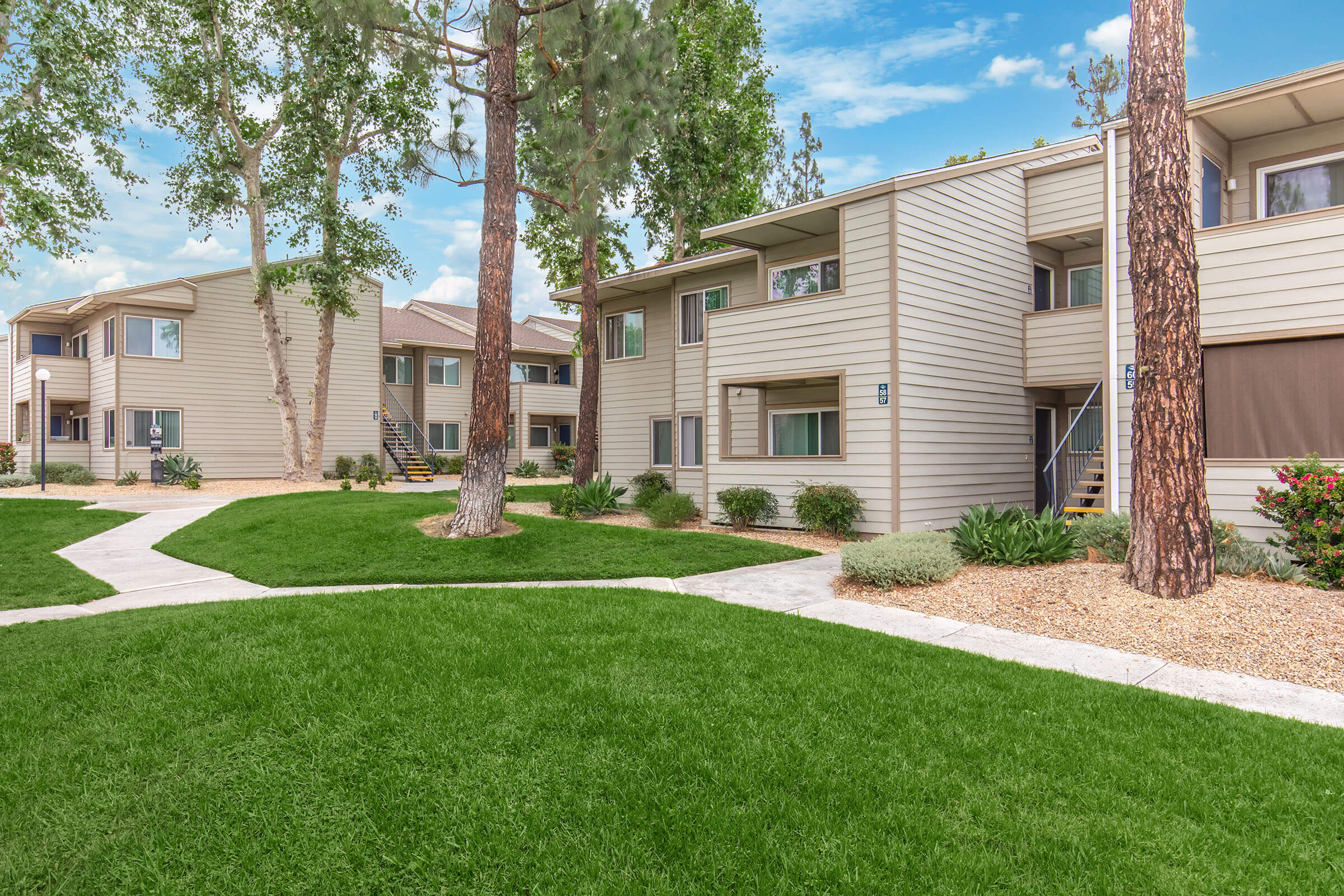 A landscaped outdoor area featuring several residential apartment buildings. The buildings are two stories tall with light-colored siding, surrounded by well-maintained grass and trees. A walkway winds through the green space, leading to the entrances of the apartments. The sky is clear and bright.