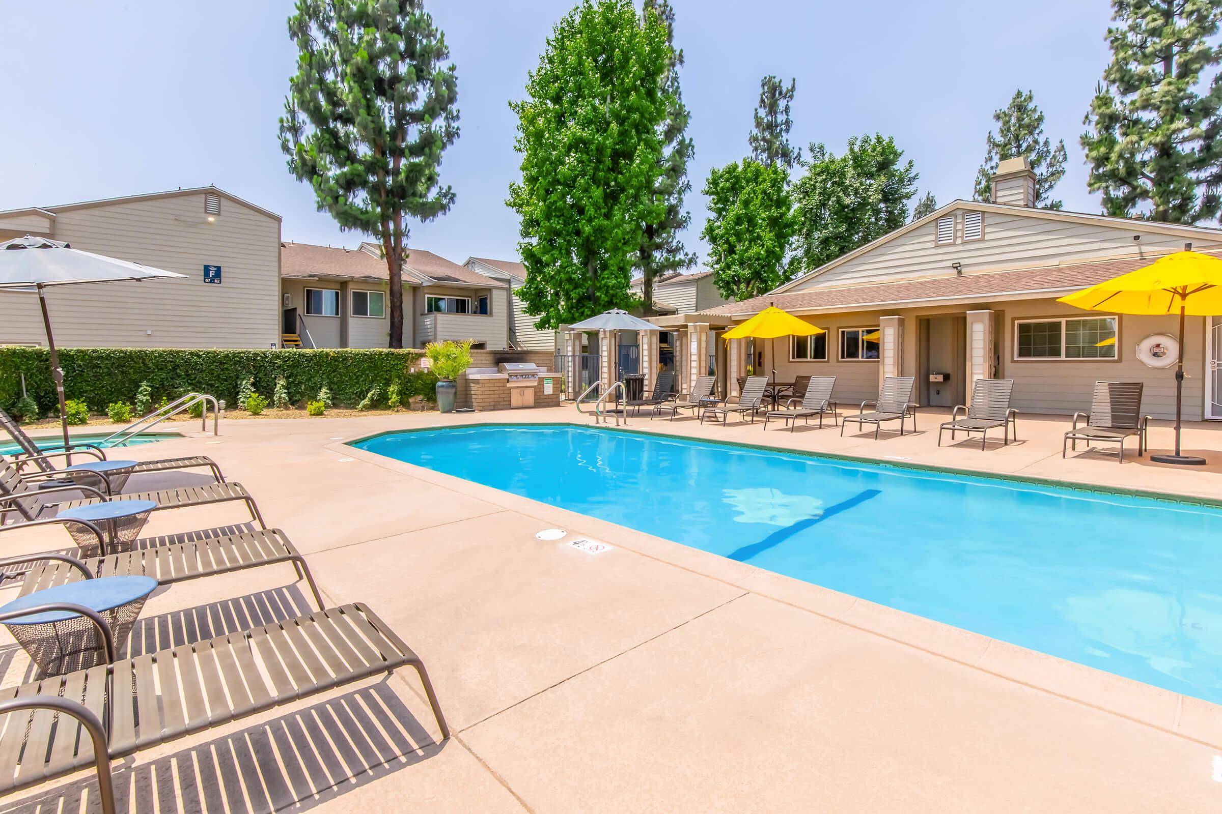 A sunny pool area with a clear blue swimming pool surrounded by lounge chairs. Yellow umbrellas provide shade, and there are trees in the background. Nearby, a building with a porch can be seen, creating a relaxing and inviting atmosphere for leisure and outdoor enjoyment.