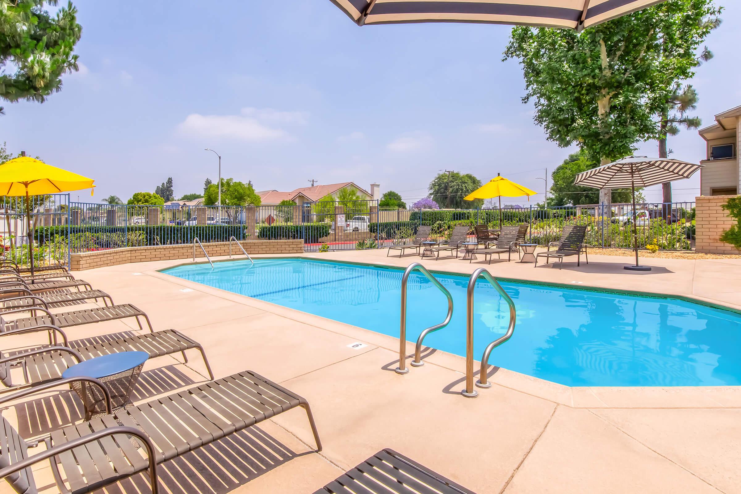 A tranquil swimming pool area featuring clear blue water, surrounded by lounge chairs, yellow umbrellas, and lush landscaping. The scene is sunny with a few clouds in the sky, creating a relaxing atmosphere for leisure and enjoyment.
