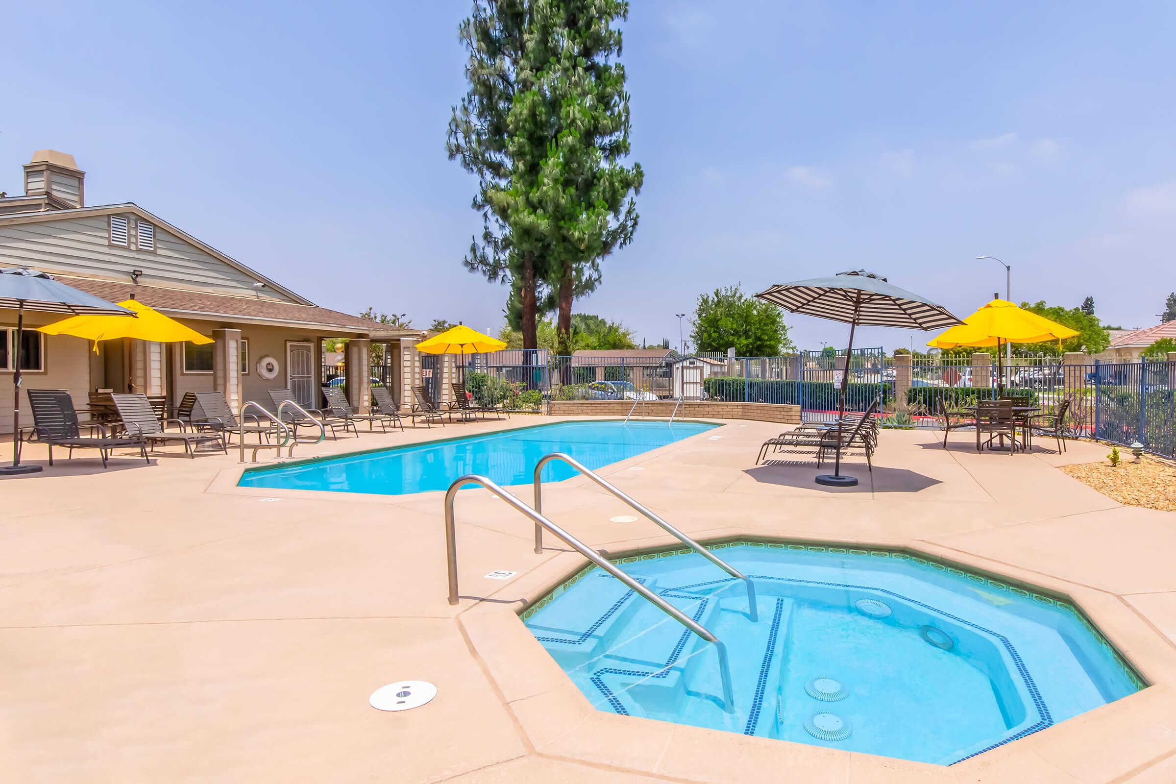 A relaxing outdoor area featuring two swimming pools, one larger and rectangular, the other smaller and octagonal, surrounded by lounge chairs and yellow umbrellas. Lush greenery and a fenced-in backyard can be seen, with clear blue skies overhead. Ideal for leisure and enjoyment.
