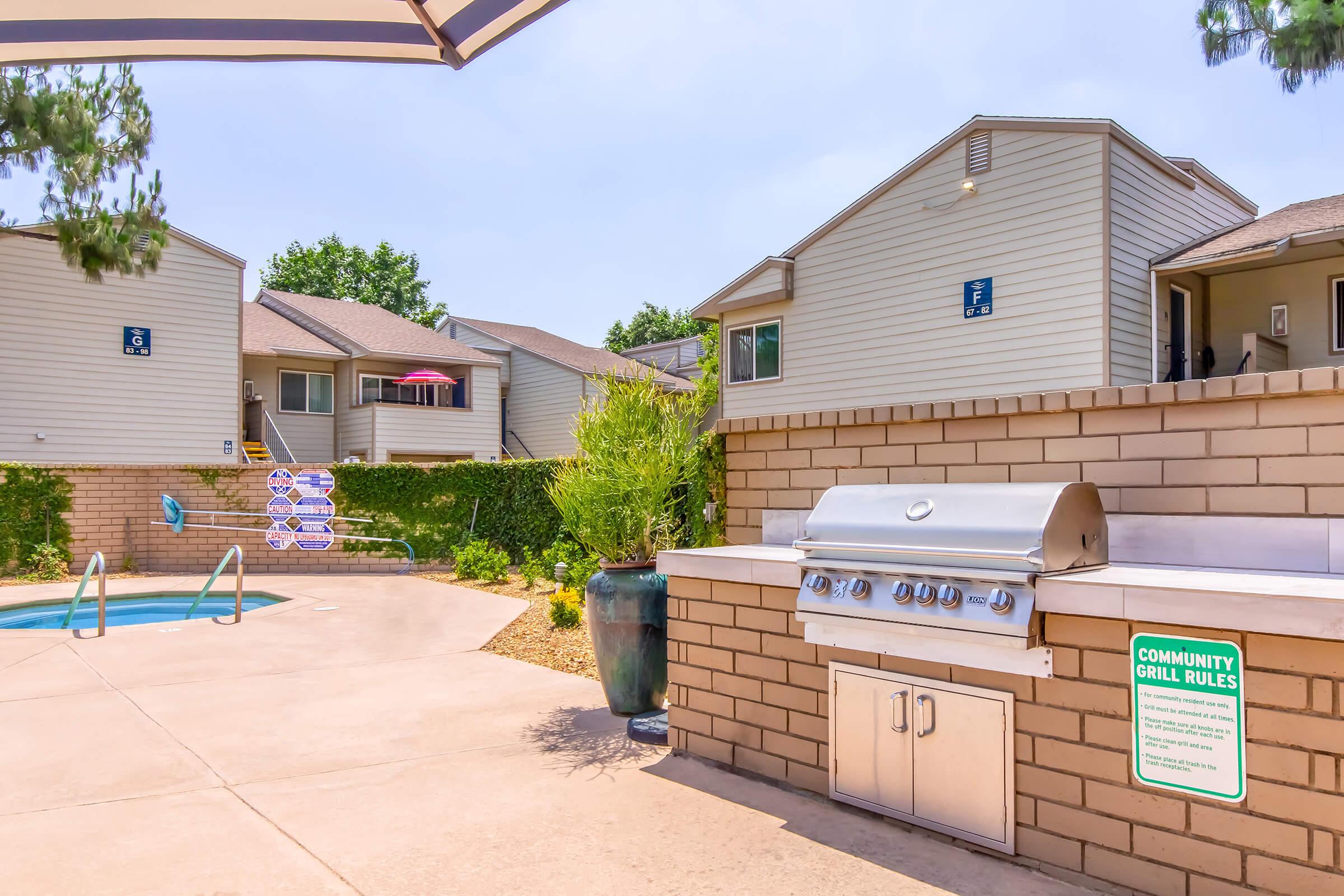 Outdoor community area featuring a barbecue grill with a sign displaying grill rules, a swimming pool in the background, and several residential buildings. Surrounding greenery and lounge chairs enhance the inviting atmosphere. Perfect for leisure and social gatherings.