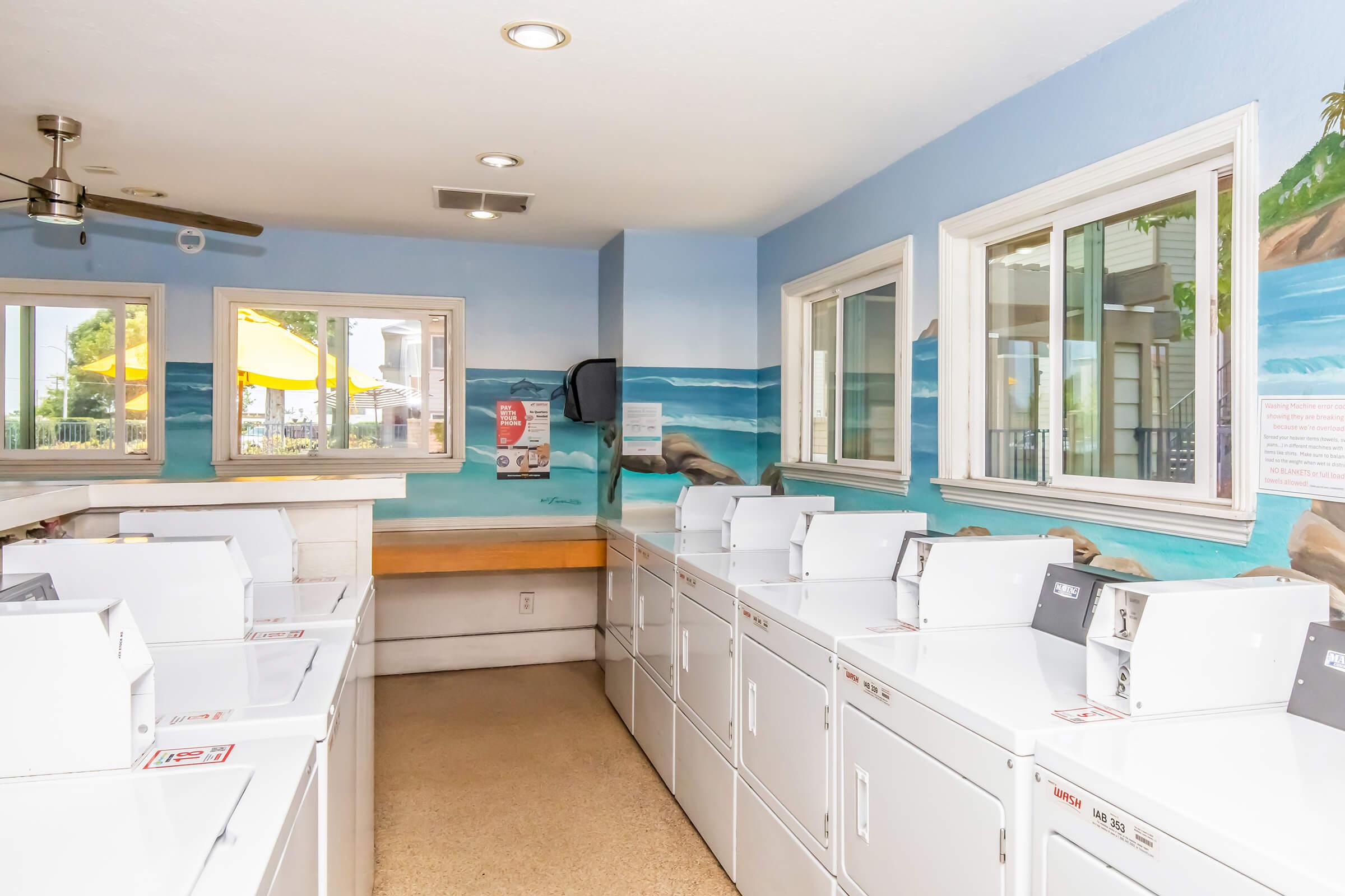 A bright, clean laundry room featuring several white washing machines lined up against a blue wall adorned with a beach mural. The room has large windows allowing natural light, a ceiling fan, and a seating area with a wooden bench along one wall.
