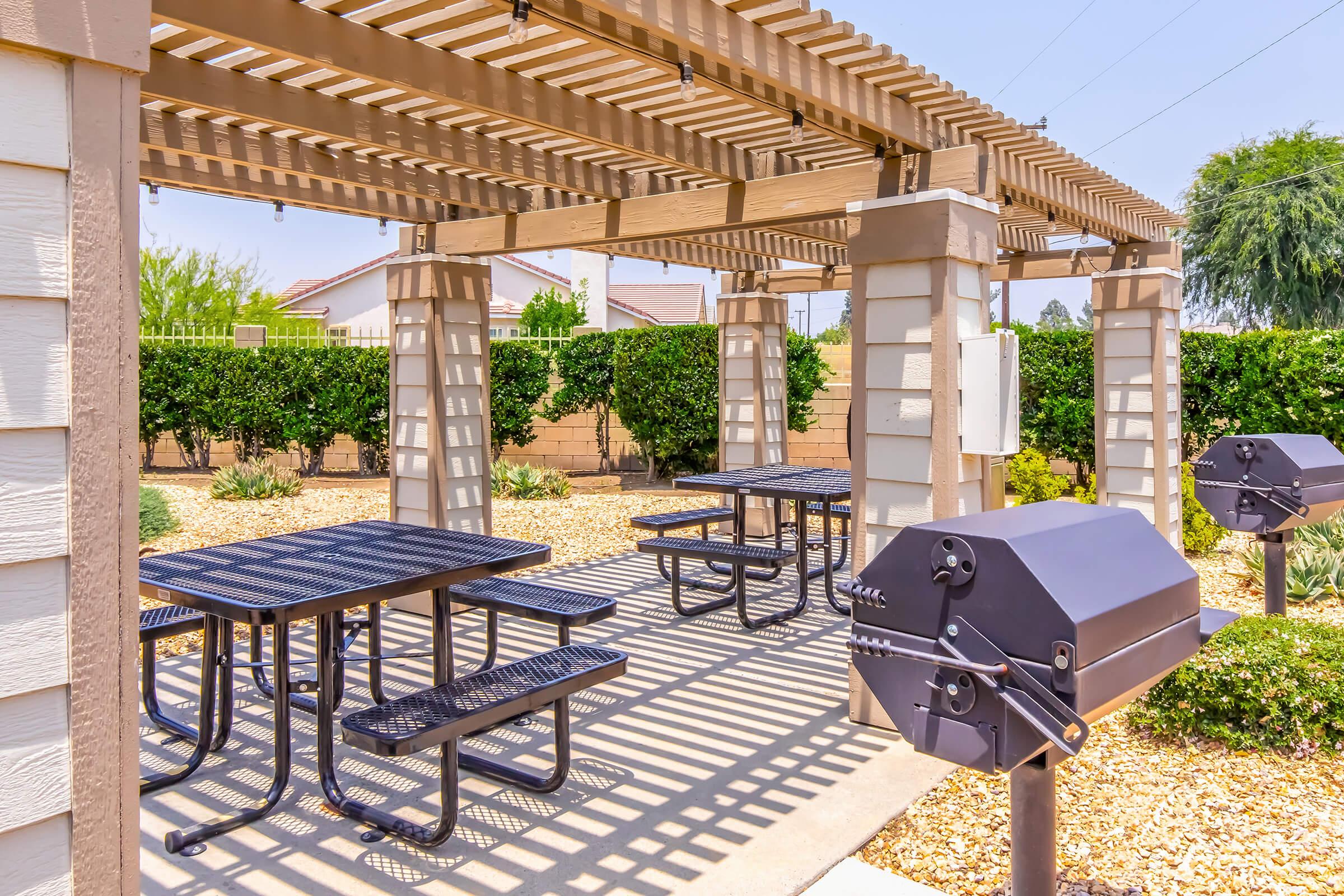 Outdoor picnic area featuring a covered pergola with multiple black picnic tables and a charcoal grill. Surrounding the space are landscaped shrubs and trees, creating a pleasant environment for gatherings and barbecues. Sunny day with clear skies and shadows cast by the structure.