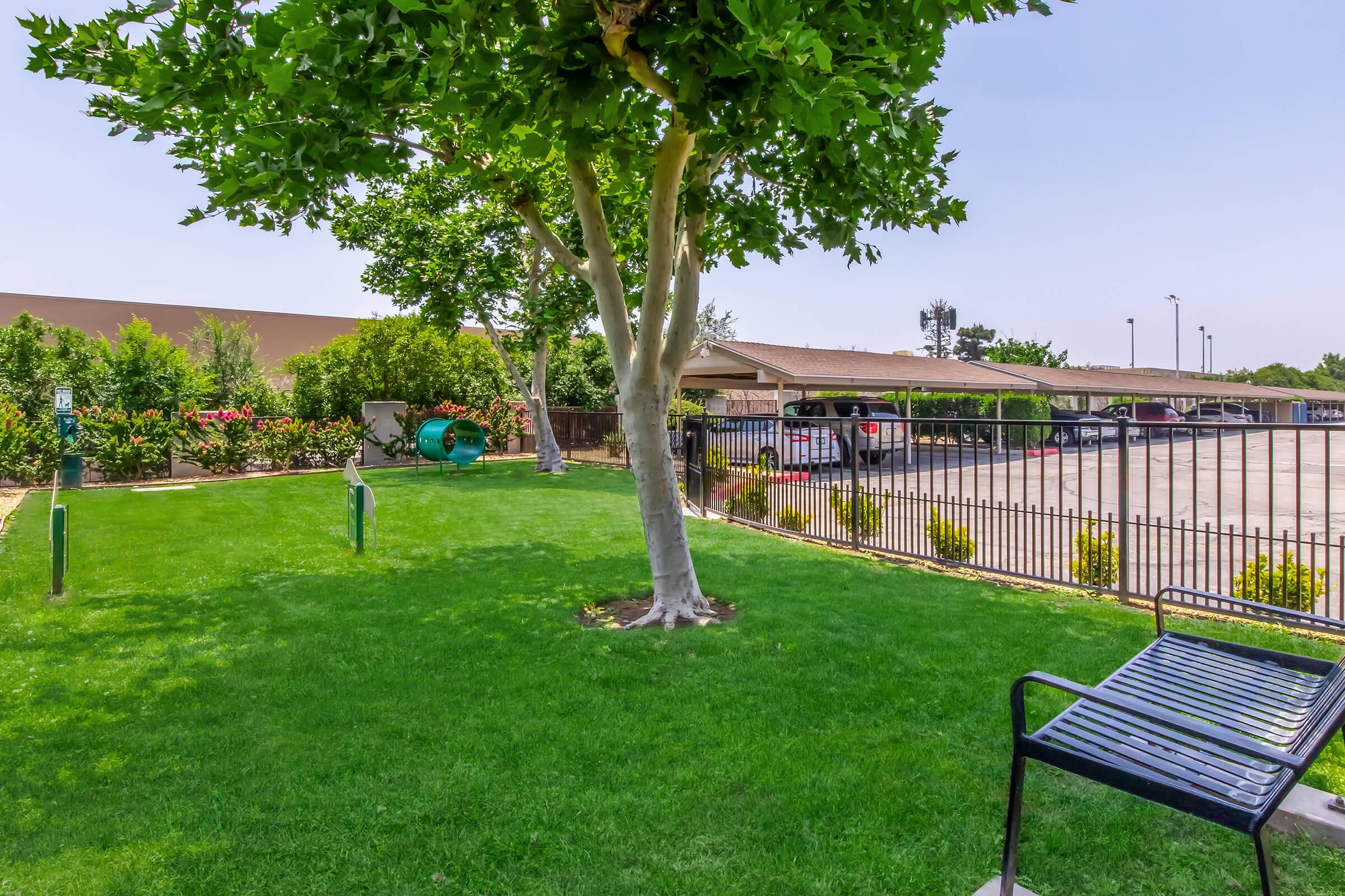 A well-maintained green lawn with a large tree, a playground area in the background, and a row of parked cars behind a black fence. Brightly colored flowers and shrubs are visible, adding to the pleasant outdoor environment. A blue bench is positioned on the lawn, inviting relaxation.
