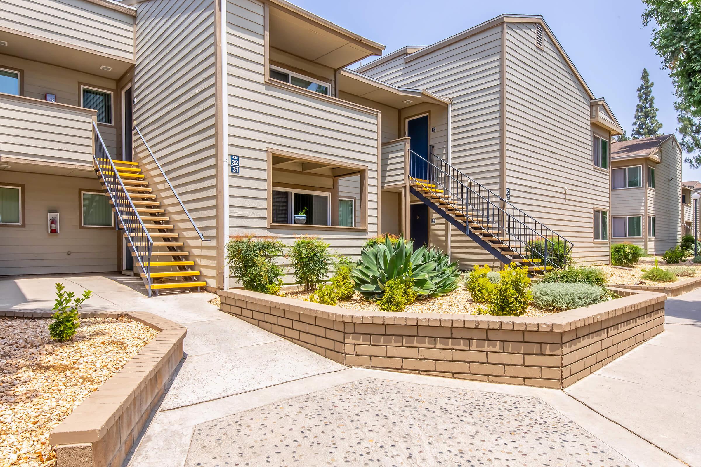 A modern apartment complex with two buildings. Staircases lead to upper-level apartments. The landscaping includes small shrubs and succulents, with a pathway made of concrete and decorative gravel. Clear blue sky overhead.